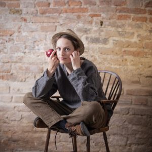 Mushroom brown linen pants with high waist, elastic waistband, and tapered legs, worn by a seated model in brown boots and grey long-sleeve top. Model holds red apple, sitting on wooden chair with grey cloth-covered floor and rustic brick wall backdrop. Keywords: bohemian pants, high waist pants, loose linen pants, linen pants women.