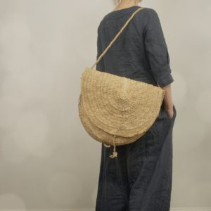 Back view of one model wearing a dark navy blue linen maxi dress with 3/4 sleeves and side pockets, kaftan style and loose fitting, standing indoors on dark wooden floor, styled with a large woven straw shoulder bag.
