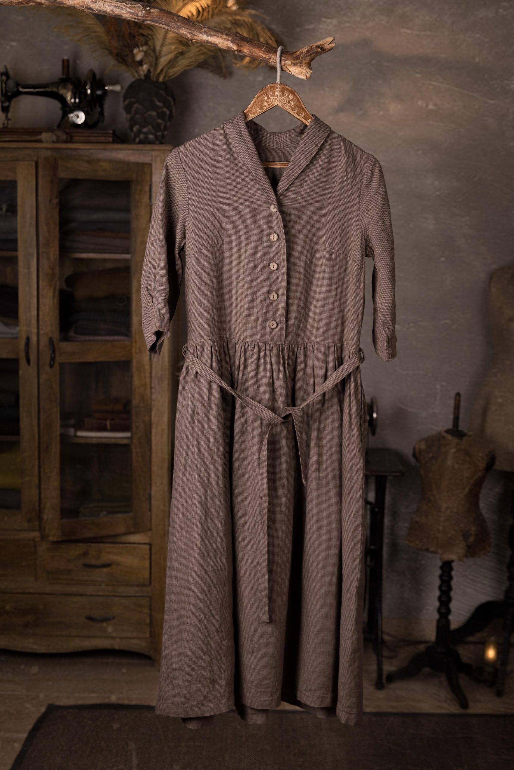 Taupe linen button down dress hanging on a wooden hanger, showing shawl V-neck collar, 3/4 sleeves, gathered skirt, and tie belt, with a rustic cabinet in the background.