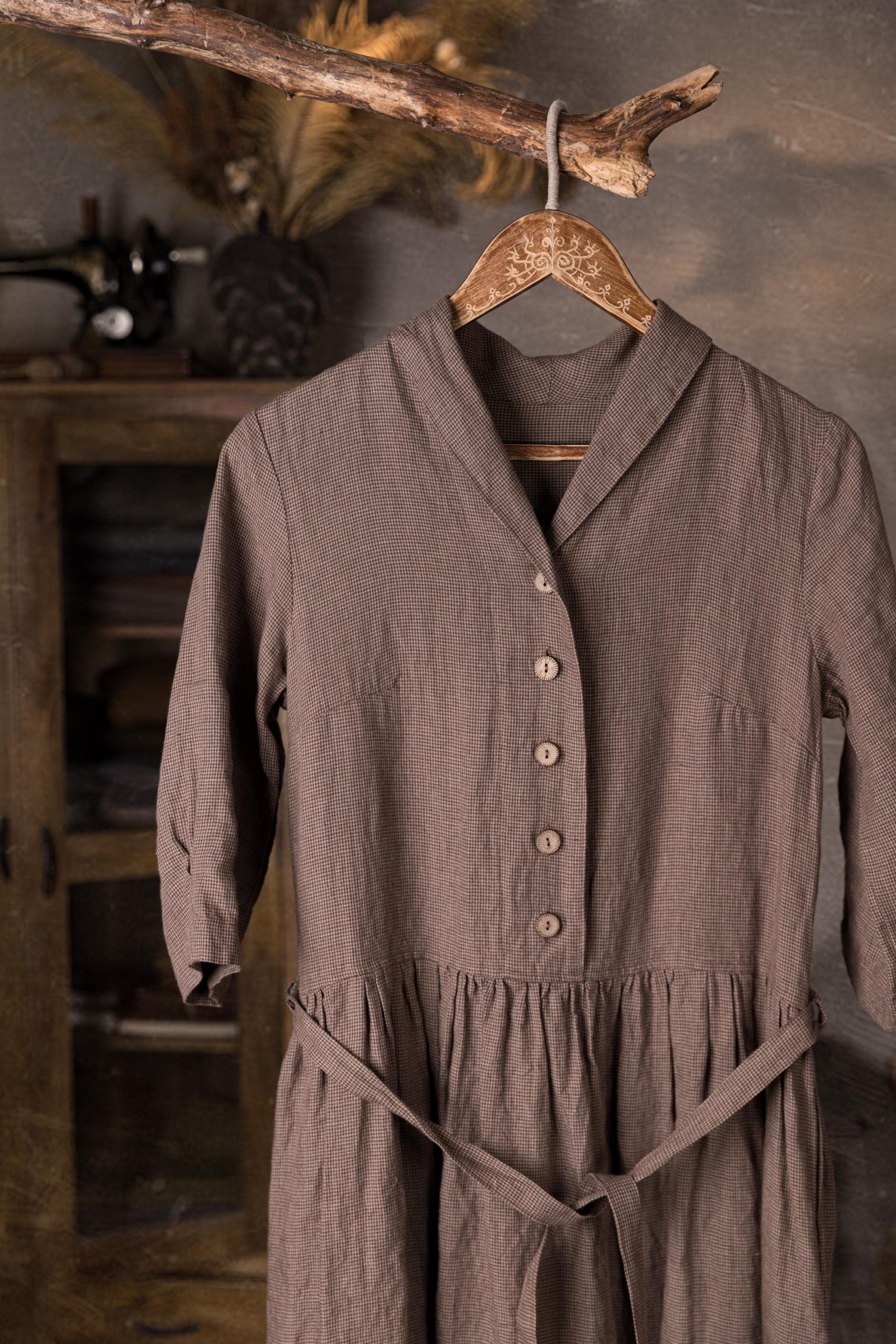 One taupe linen dress hanging on a wooden hanger, close-up of the bodice with V-neck shawl collar and front buttons, with gathered waist and tie belt visible.