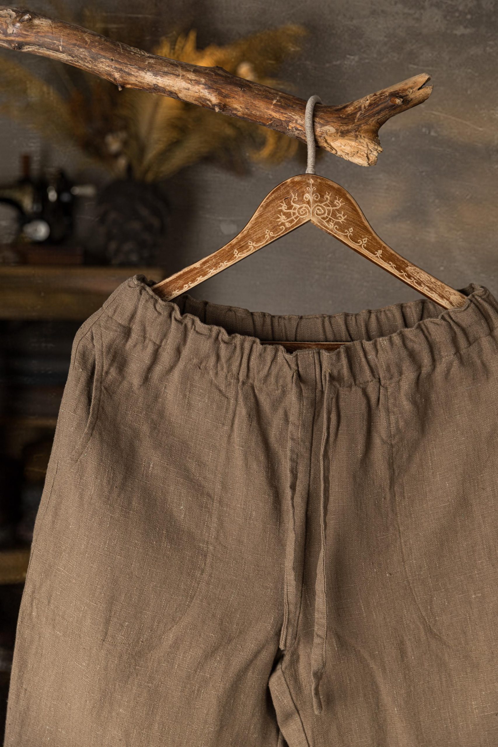 Brown linen drawstring pants hanging on hanger, waist-up front crop, ruffled elastic waistband, long narrow tie at center front, plain front with visible seam, soft creases in fabric, branch and gray wall behind.