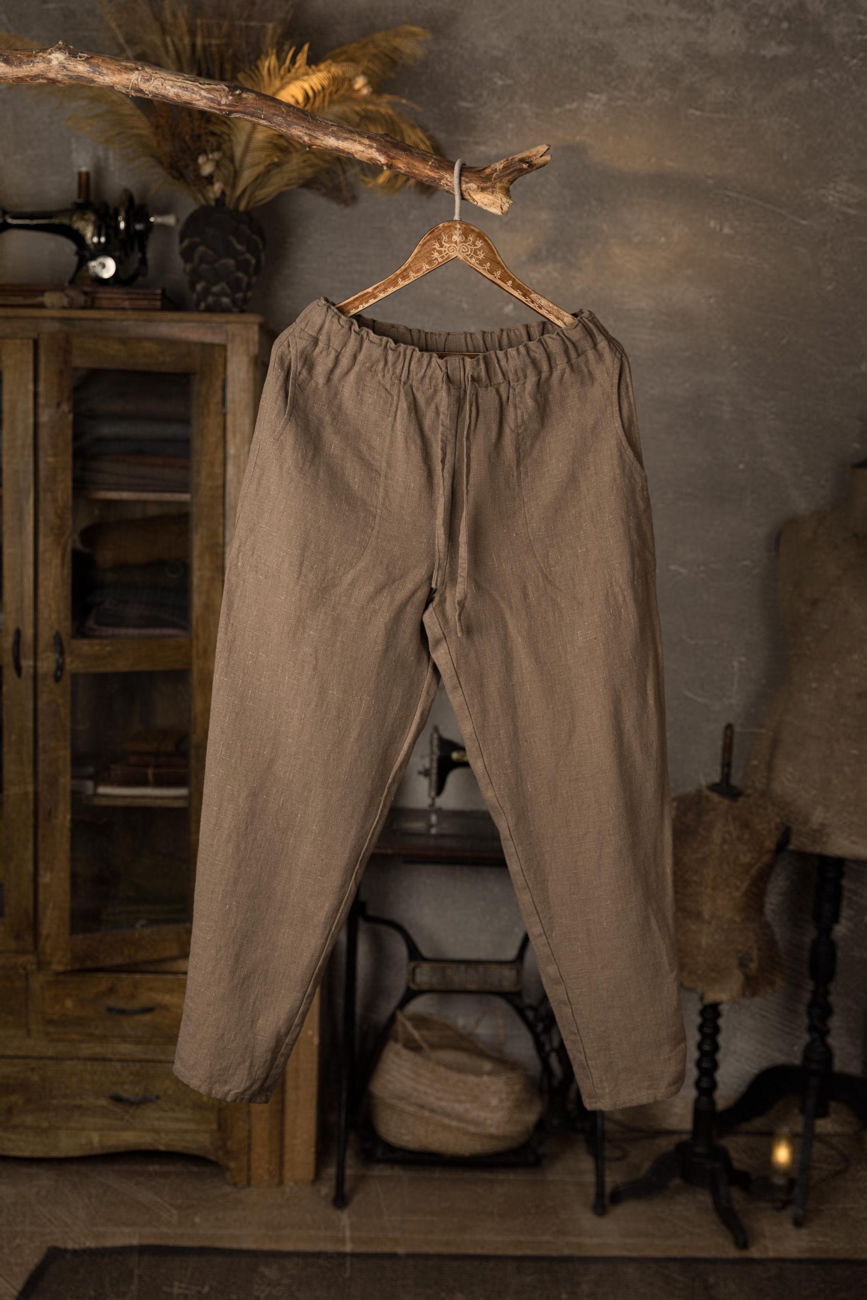 Linen pants on hanger, full-length front view, gathered waistband, loose thigh area with creases, narrower lower legs, smooth plain front panel, displayed in studio with wooden cabinet and sewing machine base in background.