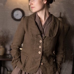 Close-up of woman wearing slim fit mushroom brown linen jacket with curved collar, button front and pockets over brown dress, shown on model in rustic studio, retro buttoned linen jacket blazer by knock knock linen.
