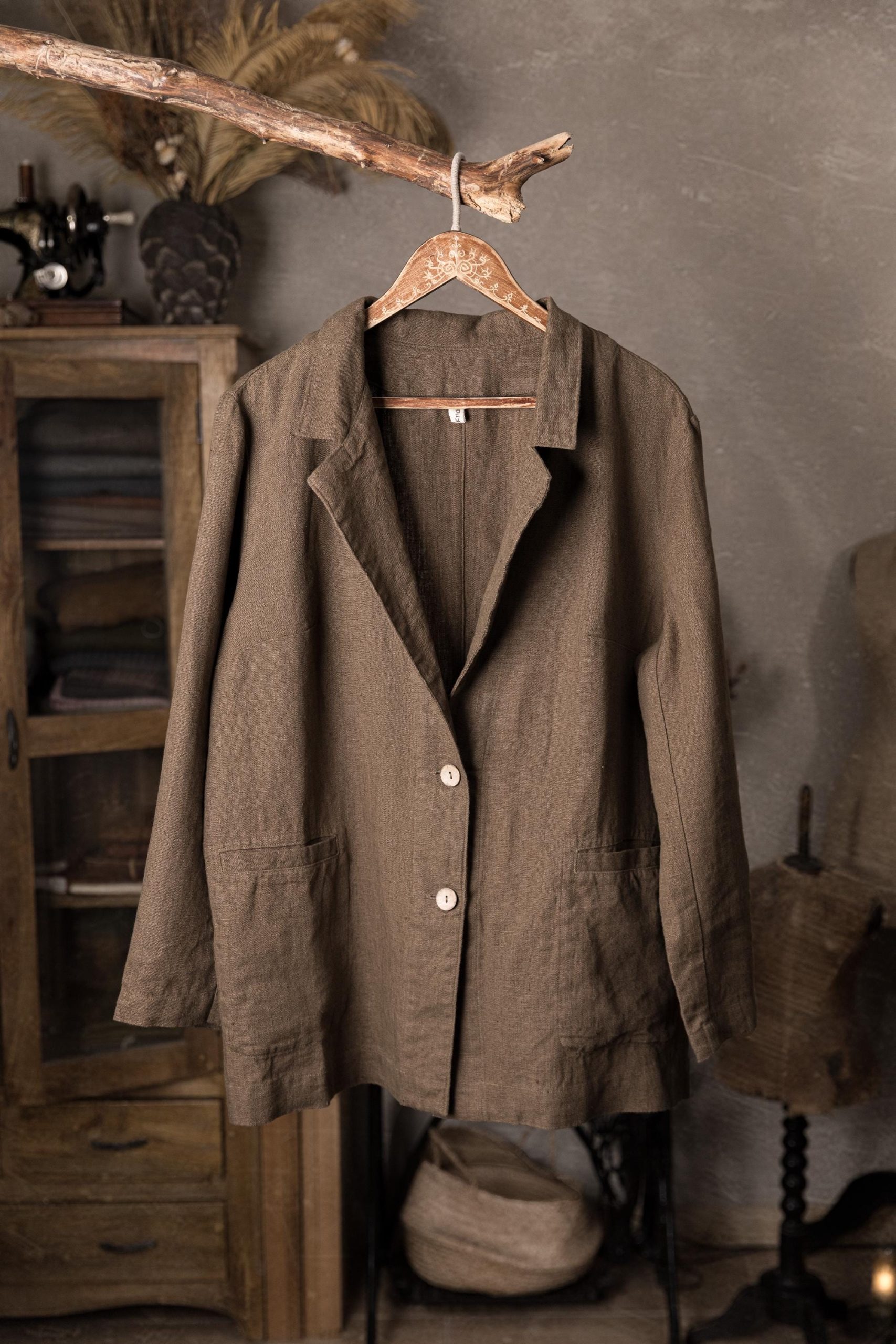 Brown linen jacket hanging on a wooden hanger, front full garment view, notched lapel collar, center front with two pale buttons, long straight sleeves, patch pockets at hip level, straight hem, rustic cabinet blurred behind.