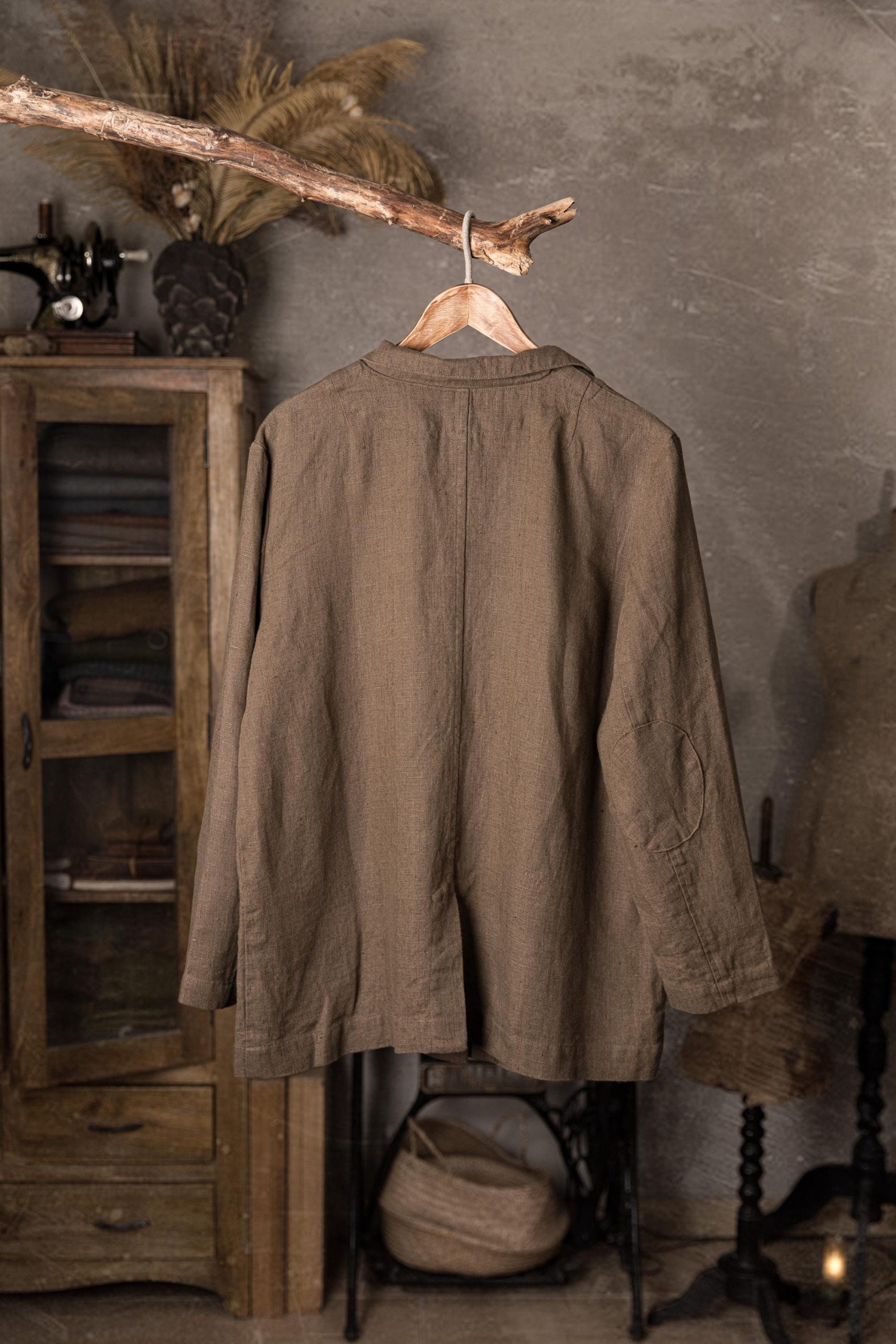 Brown linen jacket hanging on hanger from a branch, back full garment view, long sleeves with visible elbow patch, center back seam, slightly wrinkled fabric, straight hemline, rustic cabinet and dress form in background.