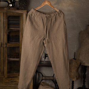 Brown linen pants hanging on a carved wooden hanger, front view full-length, featuring an elasticated gathered waistband, narrow-tapered legs, center front seam line, and a long front tie strip; sewing table in background.