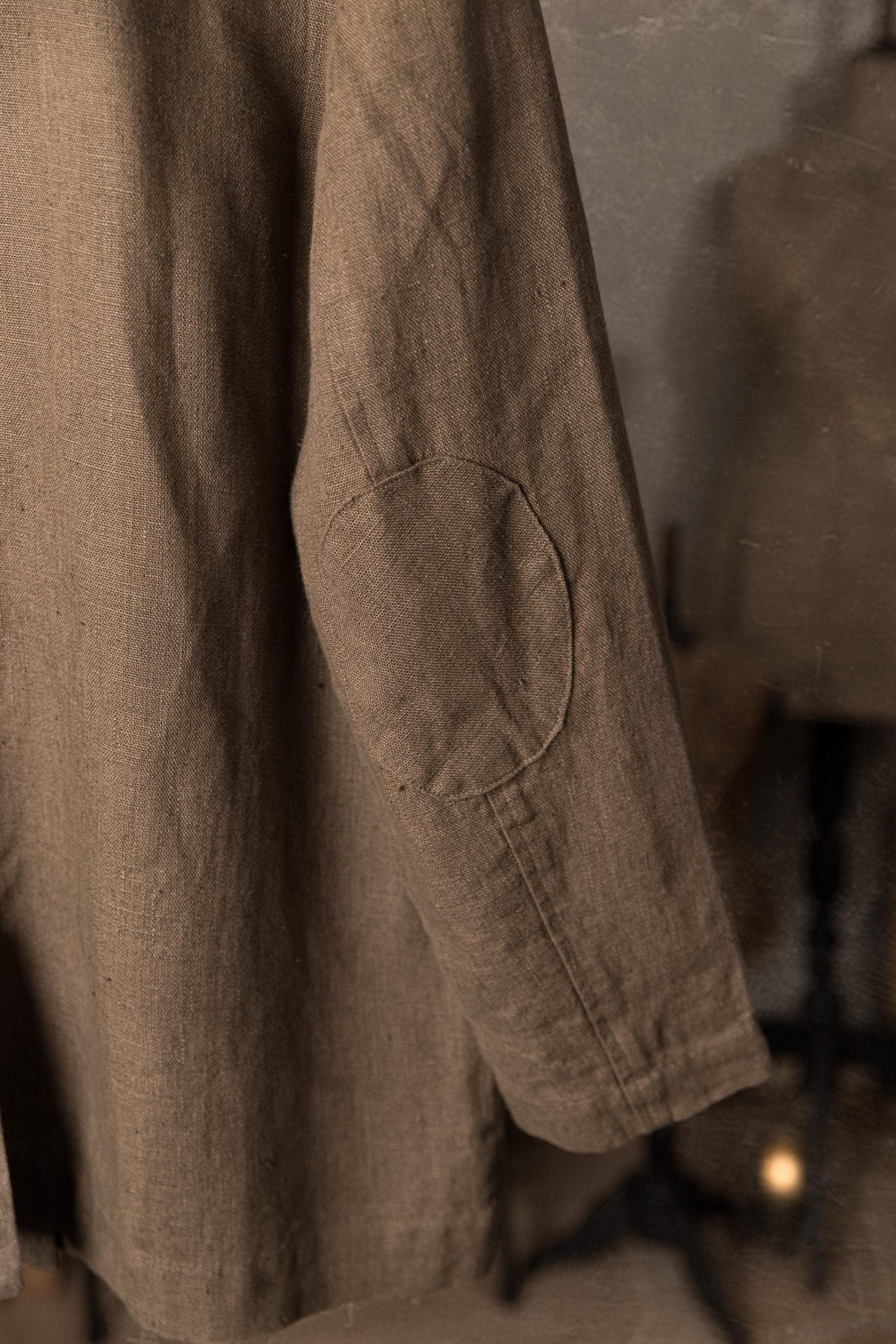 Brown linen jacket sleeve in detail close-up, centered oval elbow reinforcement patch, visible topstitching around patch, straight seam line down sleeve, wide cuff hem stitching, matte linen weave, soft-focus studio backdrop.