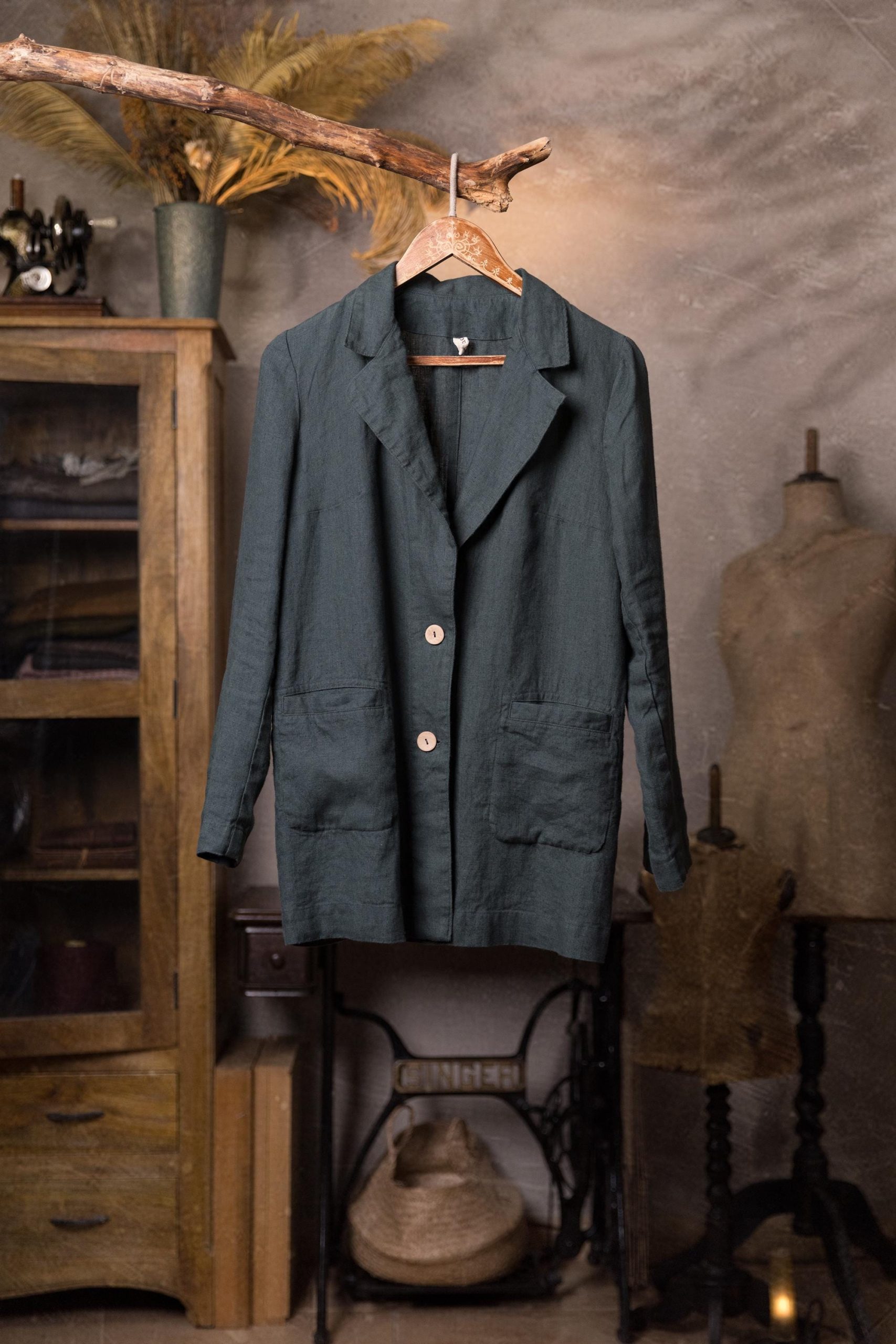 Mystic forest linen jacket on a patterned wooden hanger, full-length front view, notched collar with V opening, two light buttons, long sleeves, two large front patch pockets, hanging from a branch against gray wall.