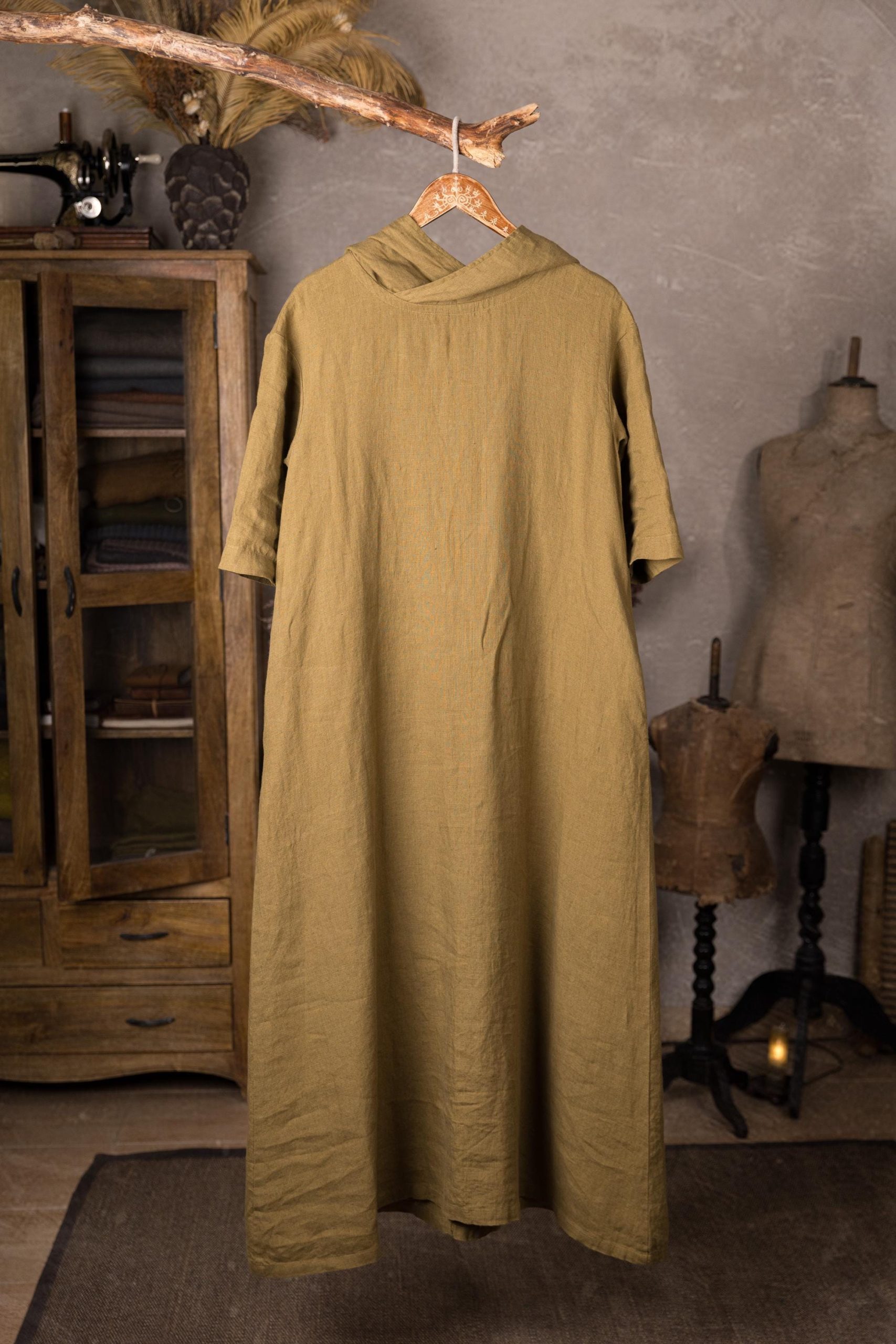 Olive green linen dress with a hood displayed full-length on a wooden hanger, back view with large hood and short sleeves, loose A-line shape, floor-length hem, photographed between a wooden cabinet and two dress forms.