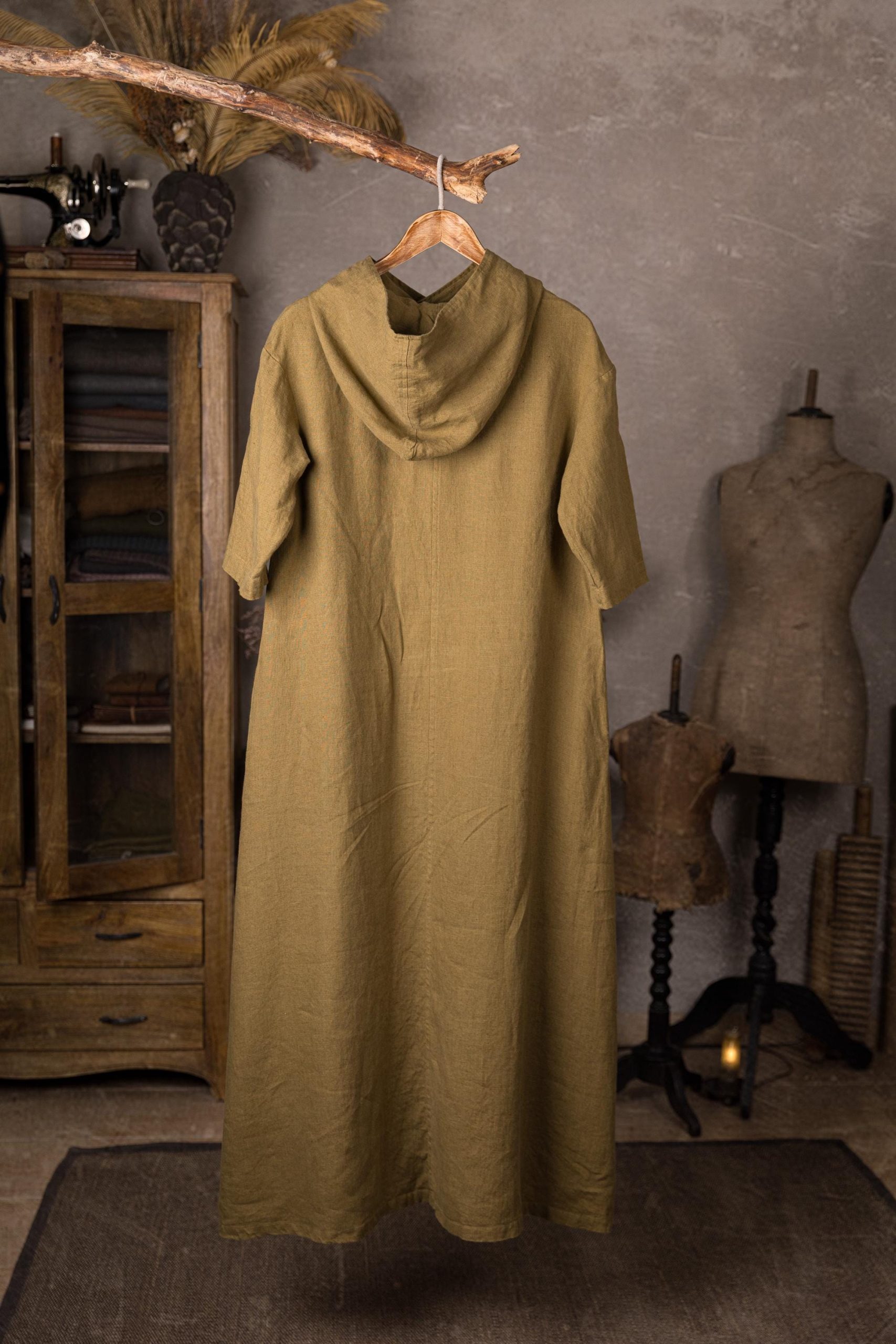 Olive linen hooded dress displayed full-length on a wooden hanger, back view with deep hood drape, center seam detail, short sleeves, plain back, soft wrinkles, rustic studio with sewing machine and glass-front cabinet.