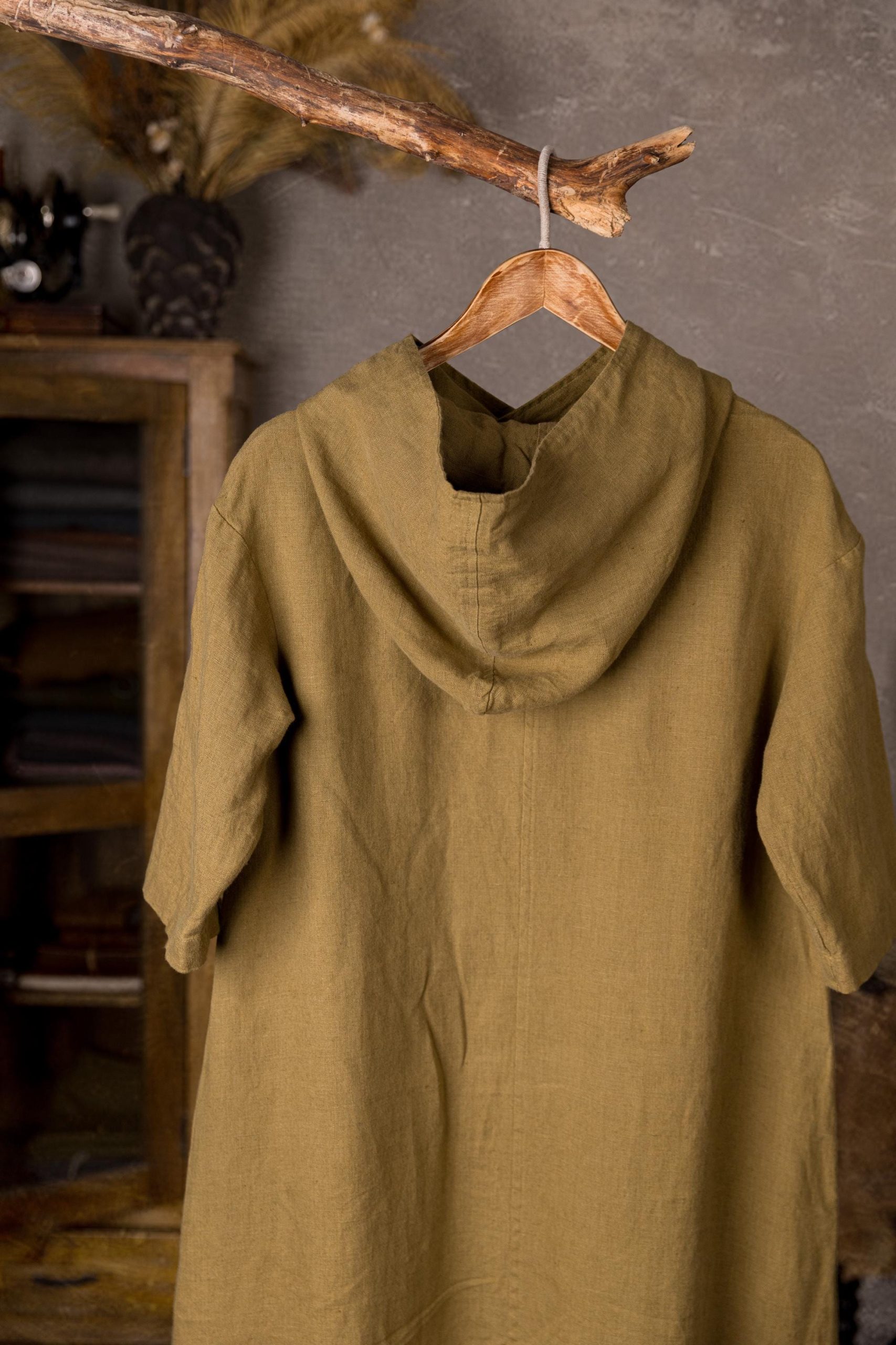 Olive green linen dress with hood displayed on a hanger, back close-up of neckline and hood, deep hood drape, center stitching, short sleeves, wrinkled linen texture, hanging from a rope on a branch, blurred cabinet and decor in background.