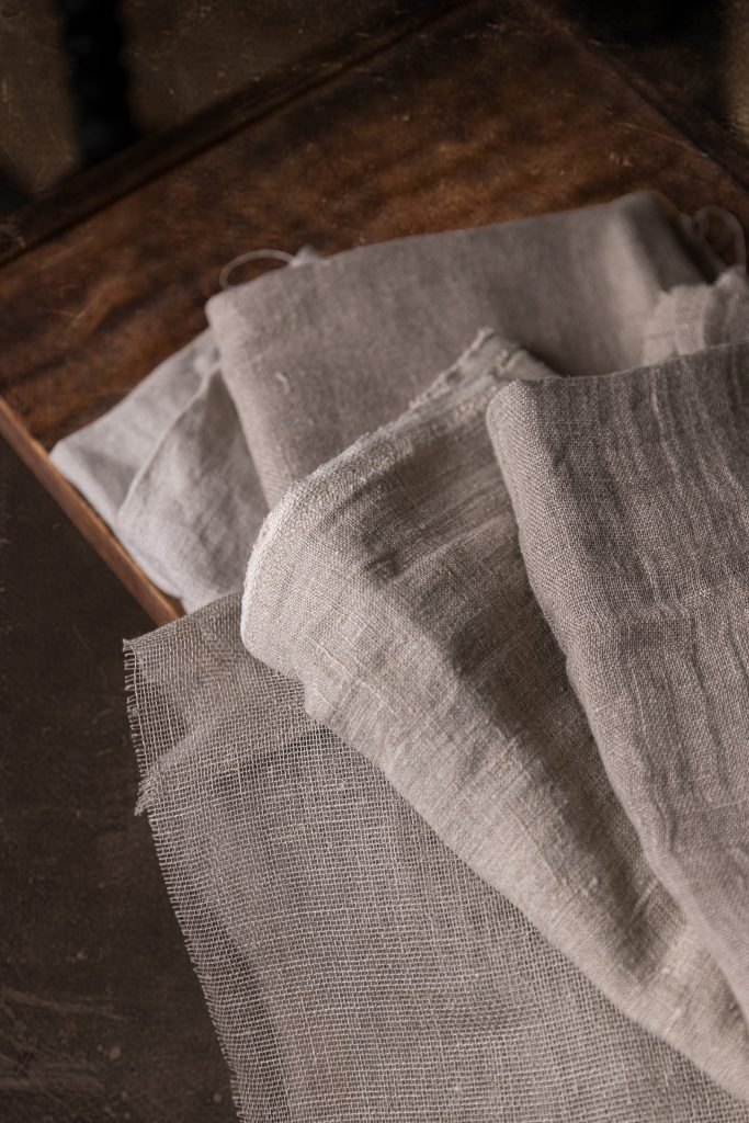 Close-up of stacked natural linen pieces in a wooden box, showing different weaves and soft creases in neutral shades.