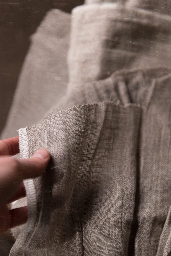 Hand holding a small piece of frayed-edge linen fabric in front of a blurred linen garment in similar natural gray-beige tones.