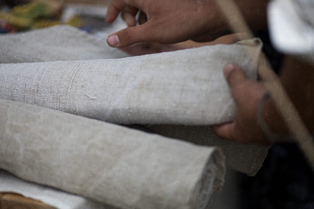 Close-up of two hands holding and lifting a roll of light gray woven fabric, with more folded fabric layers in the foreground.