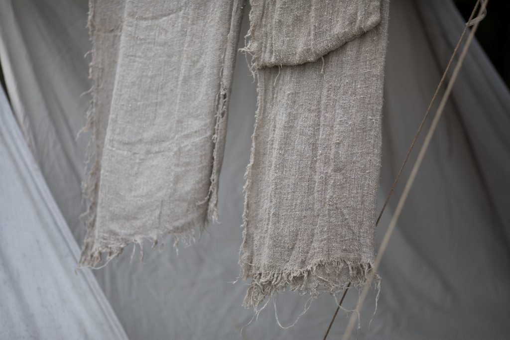 Two long pieces of light grey woven fabric hanging side by side with frayed edges, against a draped grey background