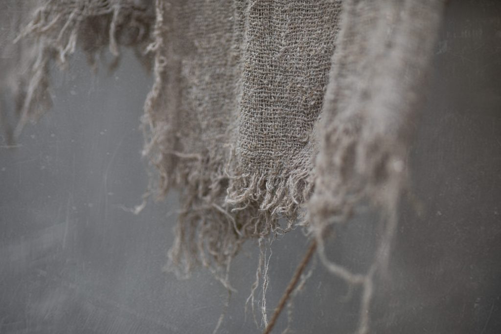 Frayed edge of coarse woven fabric draped over a thin twig, with loose fibers on a muted grey backdrop