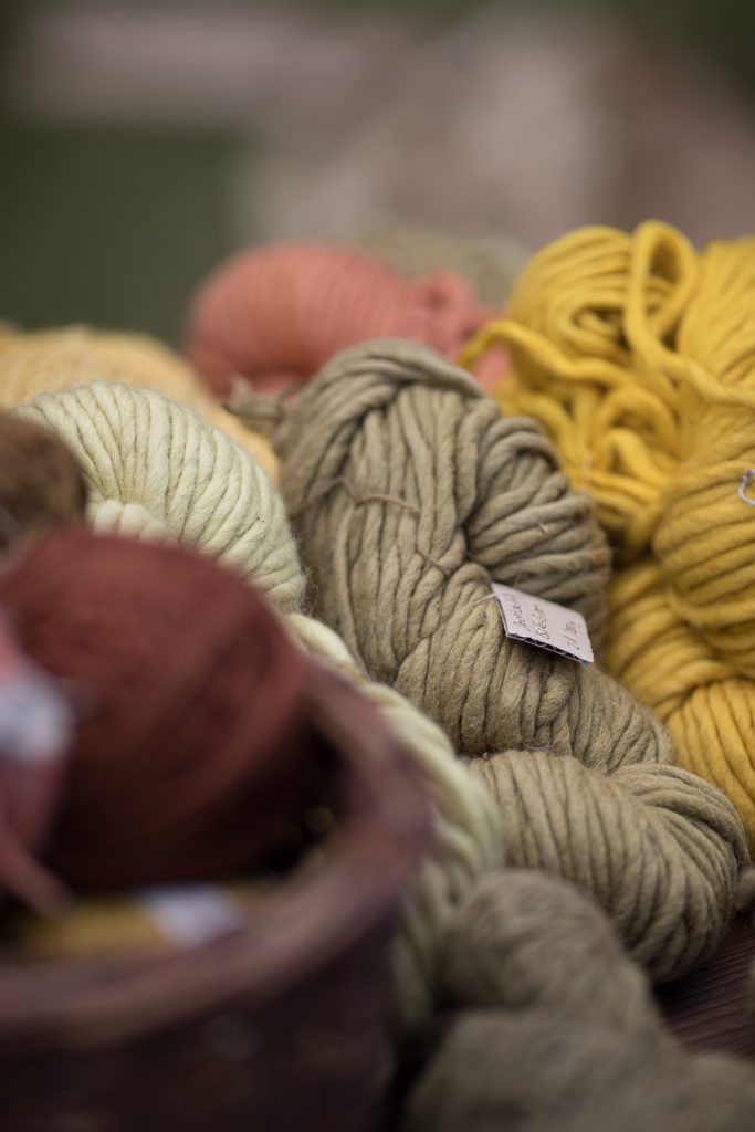 Pile of chunky yarn in mustard yellow, cream, and gray on a surface, with a blurred basket and brown yarn in the foreground.