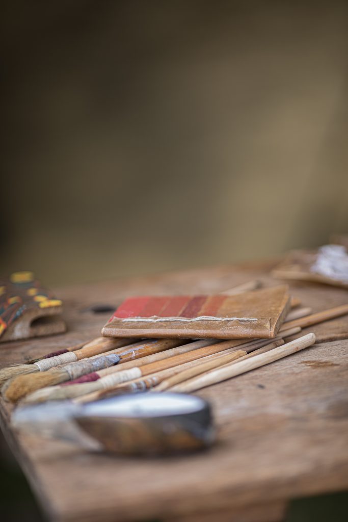 Shallow-focus view of a work table with several paintbrushes, a striped paint sample, and a blurred metal cup in the foreground.