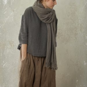 Medium grey linen/wool blend scarf with fringe, wrapped around the neck and hanging down the front on a model; side profile full-body crop shows hair in a bun, dark grey loose top, brown baggy trousers, textured grey wall. Picture taken in Knock Knock Linen Studio