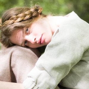 Person with braided hair wearing a natural herringbone heavy linen blouse with a boat neckline and gathered cuffs, embracing another individual in a brown garment; photographed outdoors with greenery in the background. Linen top shown on model.