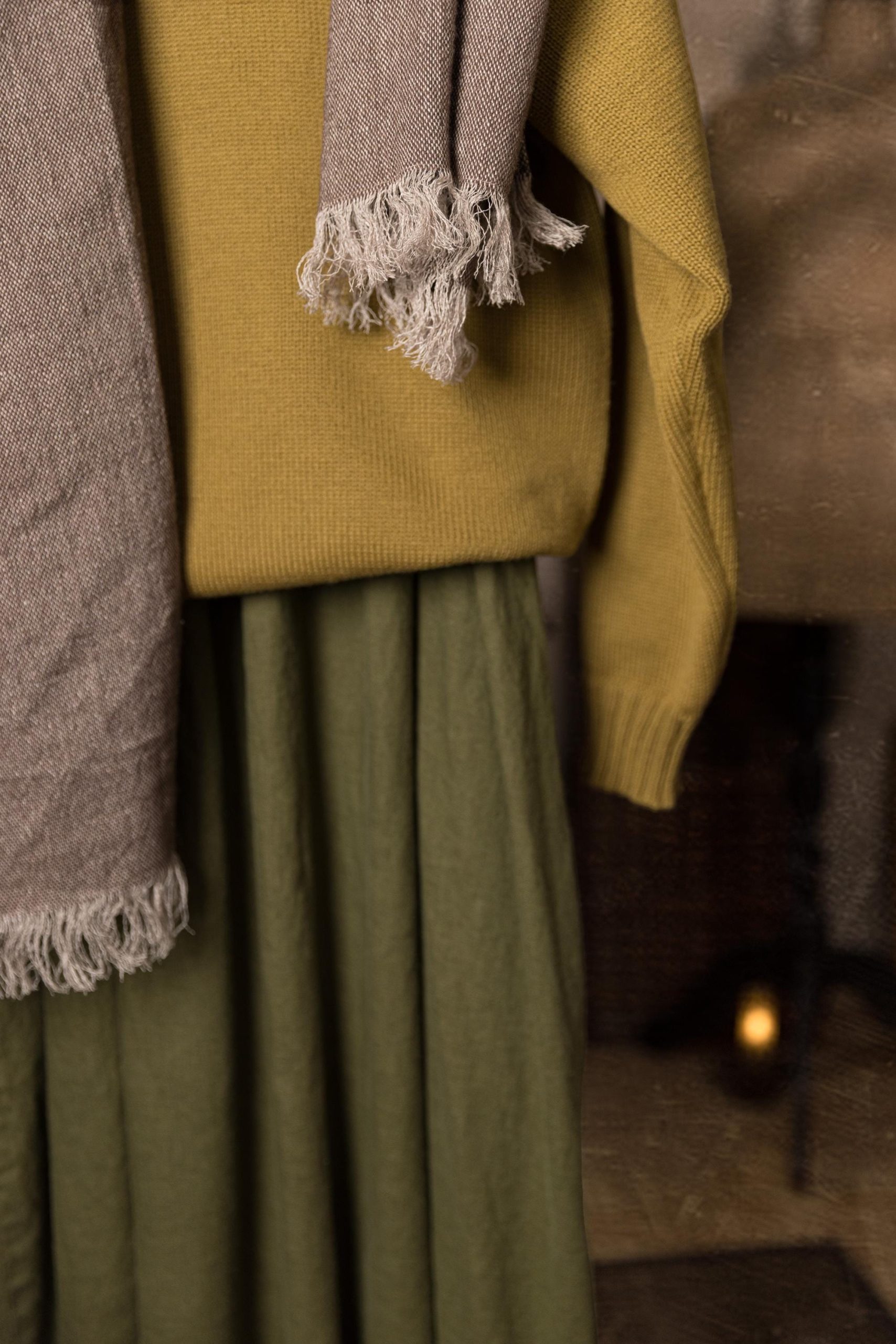 Woven scarf with pale fringe, draped on a mannequin over a mustard knit sweater; one short end hangs at center with tassels, a long panel falls down the left side; cropped front view from waist to lower skirt.