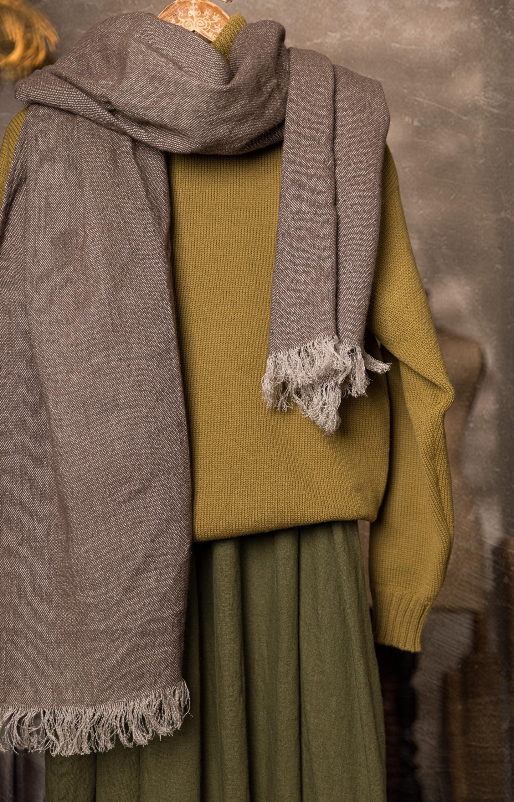 Woven scarf shown on a mannequin from chest to hips, looped once around the neck with both ends hanging; short end on the right shows loose fringe, long end falls along the left side over a mustard sweater.