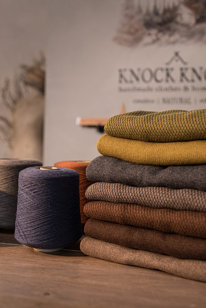 Stack of folded textiles in olive, mustard, gray, and brown tones on a wooden table, with a dark blue yarn cone beside them and a blurred “KNOCK KNOCK” sign in the background.
