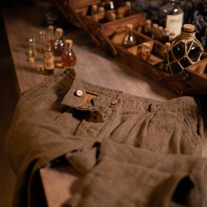 Brown linen pants in a flat lay, waist-to-thigh crop; waistband with belt loops, front pleats, tan button at fly, overlapping tab with a white button, slanted side pocket opening, and stitched seams; small bottles behind.
