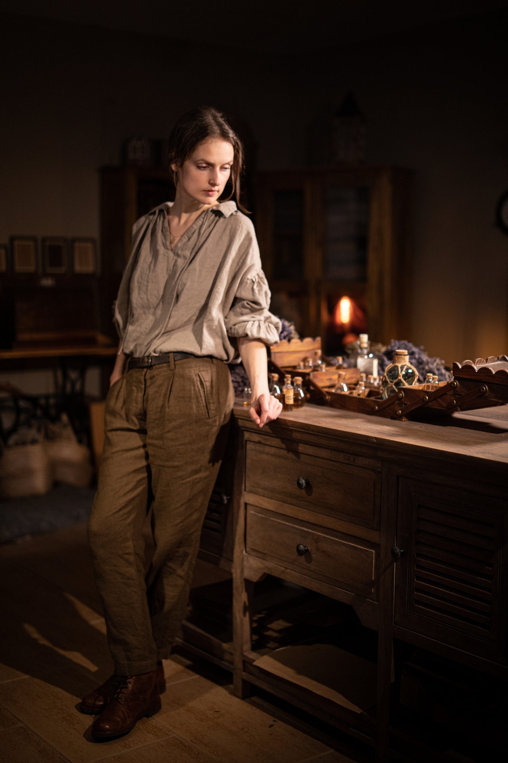 Brown linen trousers on a model, full-length three-quarter front; waistband with belt loops, pleats at the front, side pocket openings, straight fly line, legs narrowing to the ankle, and turned-up hems; dark room, bottles on counter.