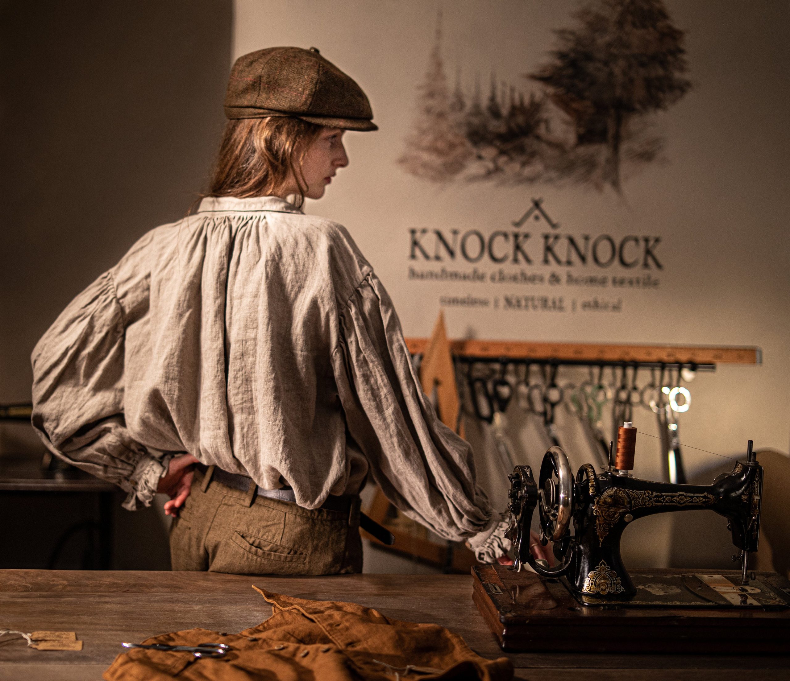 Brown linen pants on a model, three-quarter back crop; waistband and belt loops, dark belt, back pocket with button closure, angled pocket opening, creased fabric texture, model beside an ornate sewing machine; logo on wall.