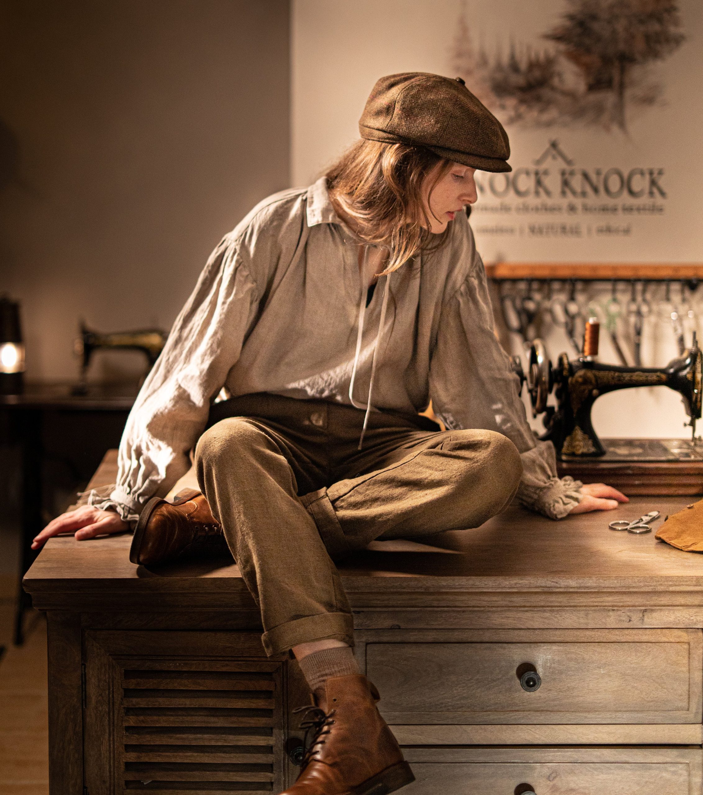 Brown linen pants worn by a seated model, full-length front view; high waistband, belt loops, front pleats, cuffed hems, one leg crossed, beige socks showing, and brown lace-up boots; vintage sewing machine on table.
