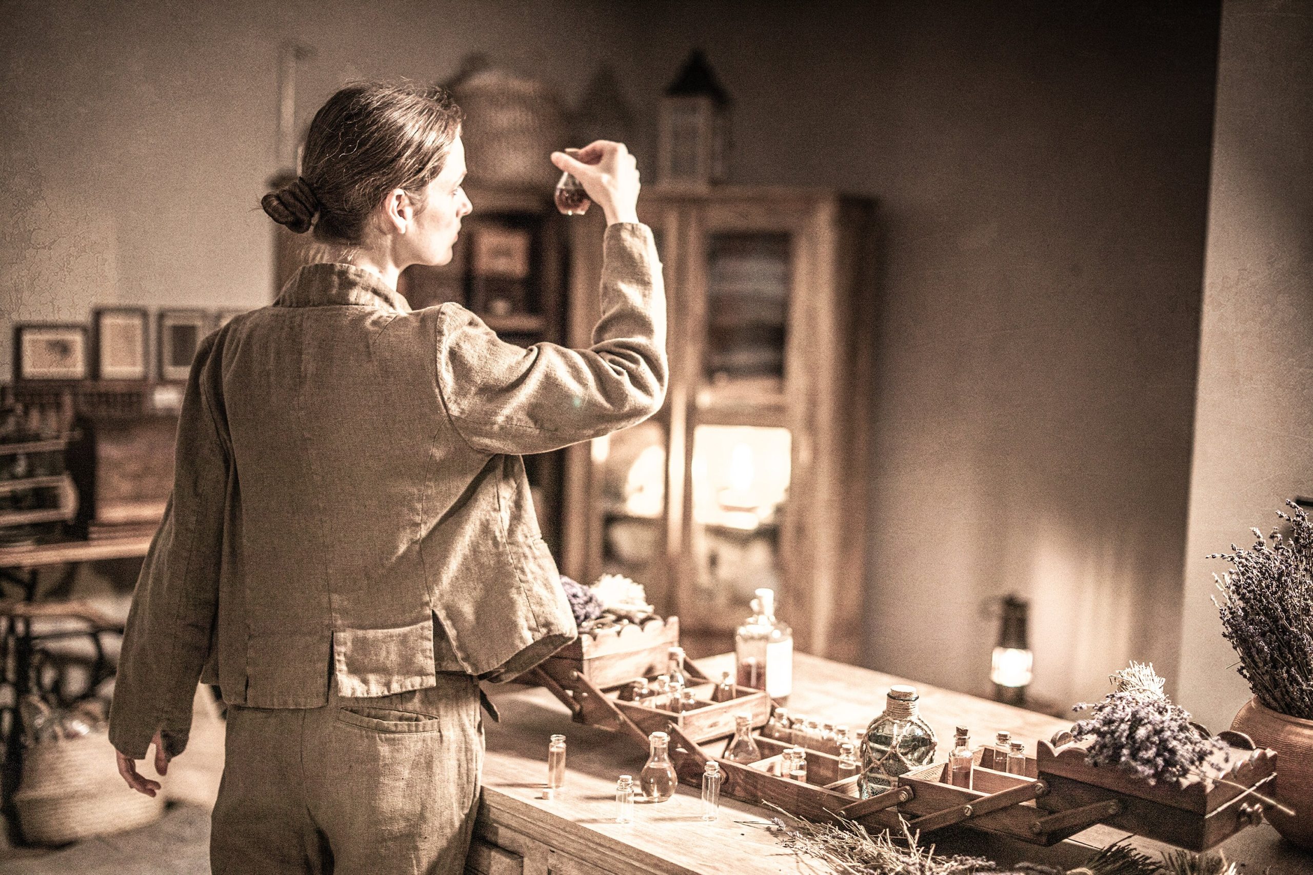 Brown linen jacket worn on a model, rear mid-thigh crop; long sleeves, straight back panel, cropped hem, seam lines at armholes, rectangular patch detail near hem, creased linen texture; wooden table with tiny bottles and dried flowers.