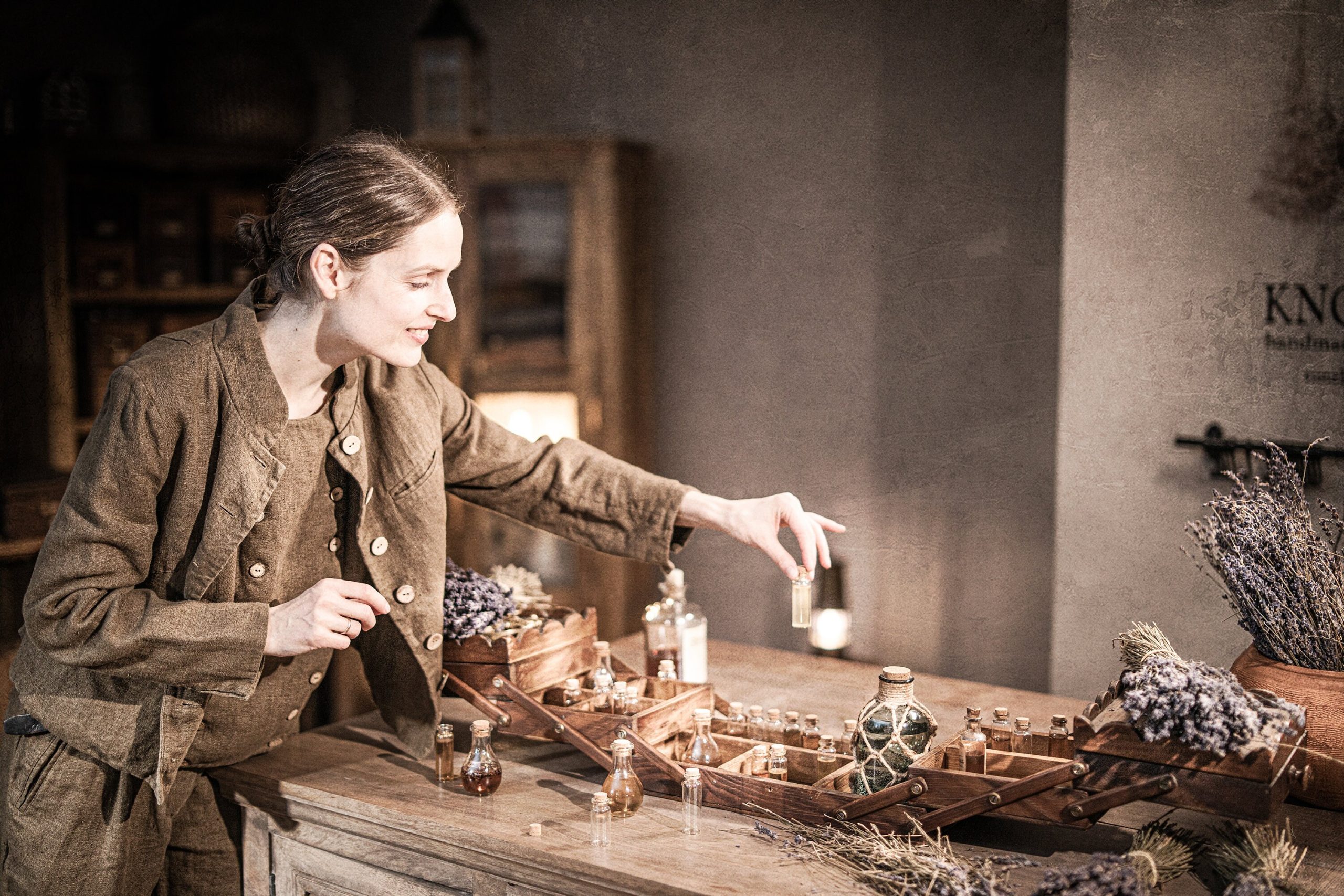 Brown linen jacket worn by a model leaning over a wooden table, waist-up side/three-quarter; light buttons down front, long sleeves, visible front seams, pocket opening at hip, and rumpled fabric; wooden tray of glass bottles and dried flowers in front.