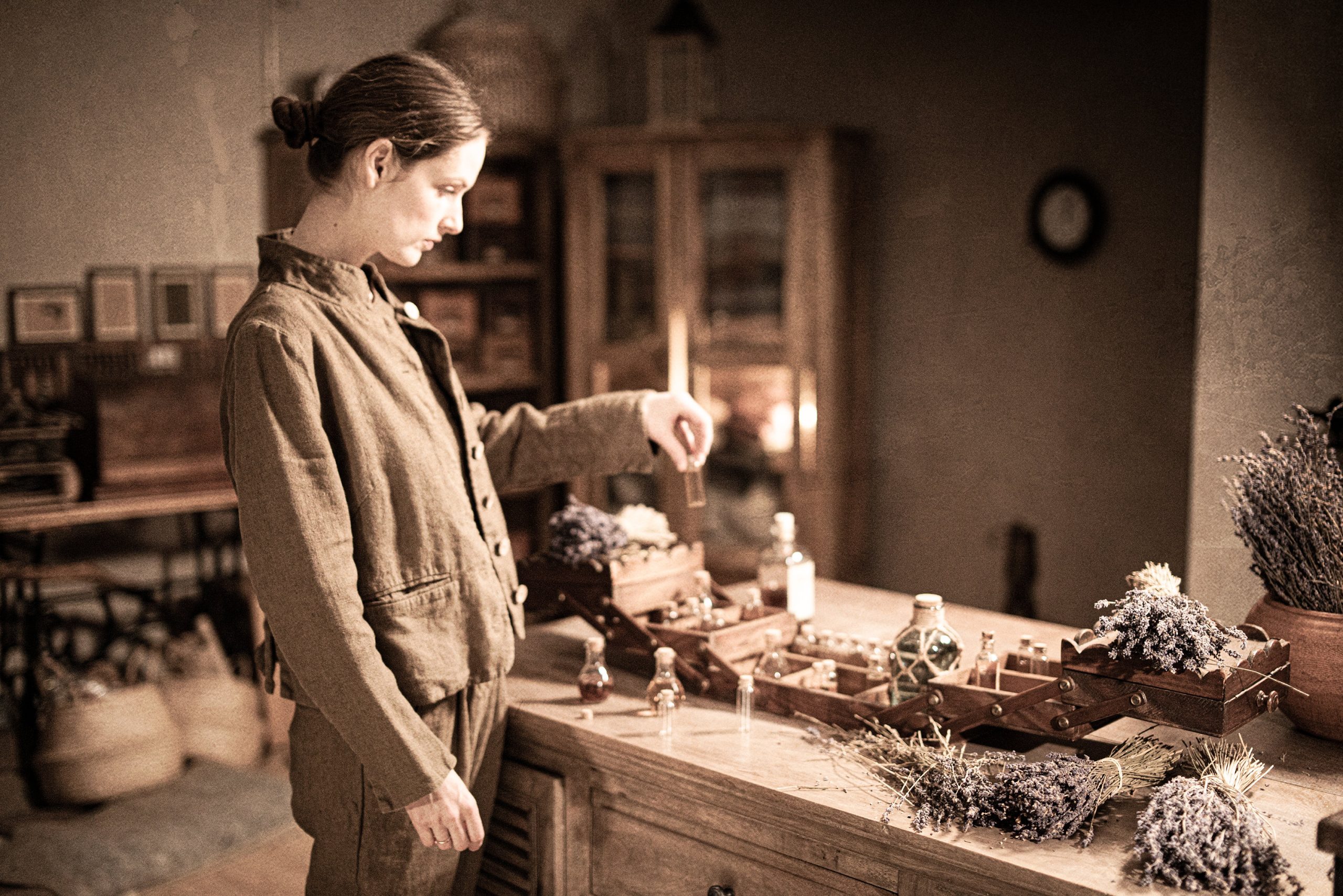 Brown linen jacket on a model, side/front three-quarter view (mid-thigh crop); stand collar, button placket, long sleeves, welt pocket at hip, straight cropped hem, and wrinkled linen surface; she looks down at a small vial beside a wooden tray of bottles.