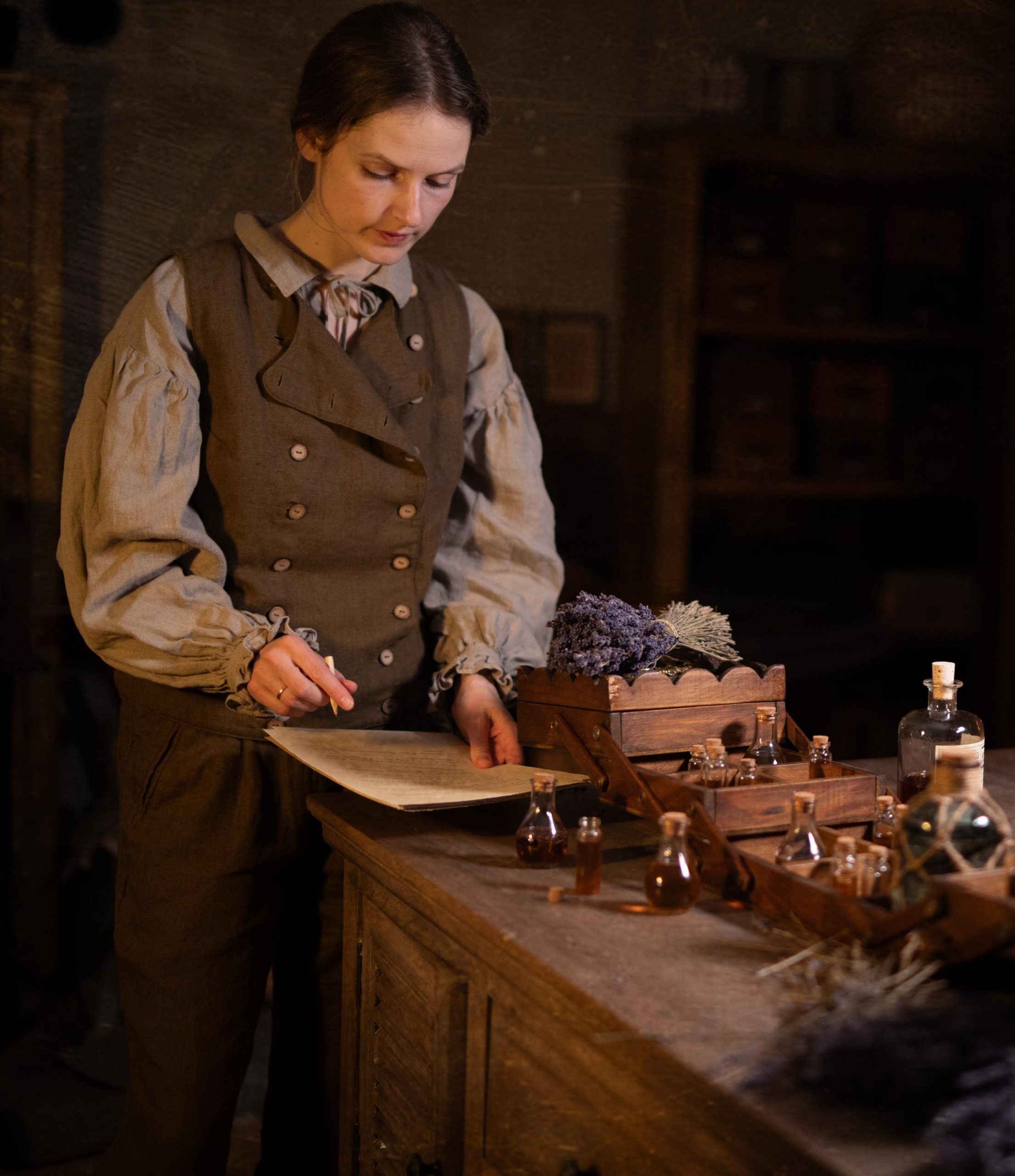 Brown linen waistcoat on model, waist-up view; sleeveless with two rows of light buttons and curved overlap; worn over pale blouse with gathered sleeves and ruffled cuffs; pencil and paper on counter with corked vials and lavender.