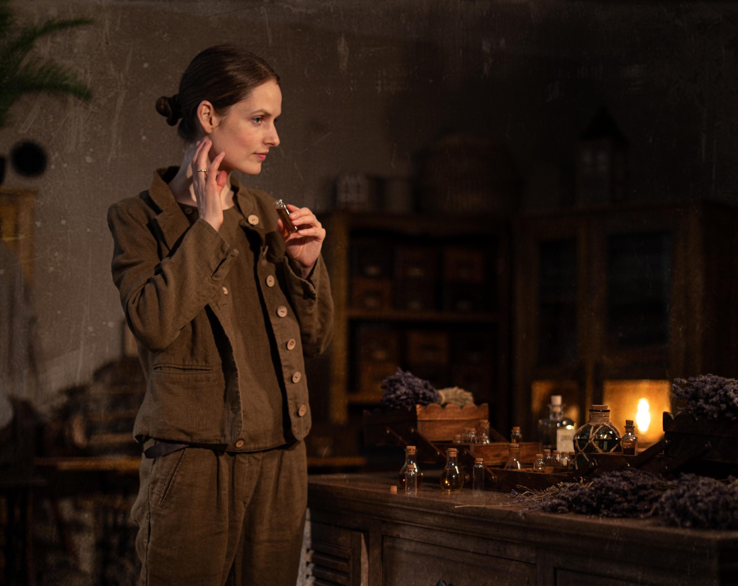 Brown linen jacket worn by a model, waist-up side view; pointed collar, row of light buttons down front, long sleeves, welt pocket at hip, front edge overlap, and linen creases; wooden table with small bottles behind.