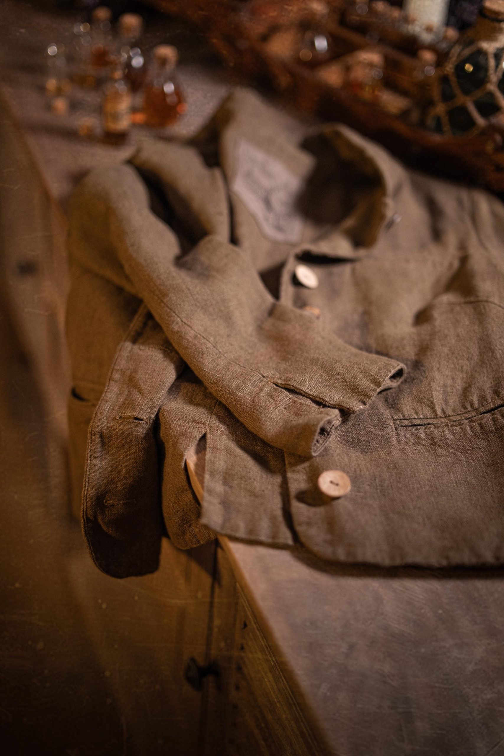 Brown linen jacket in a flat lay on a tabletop, detail view; collar area and front placket, one large light button, folded sleeve, visible welt pocket seam, stitched buttonholes, and creased fabric; small bottles out of focus at top.