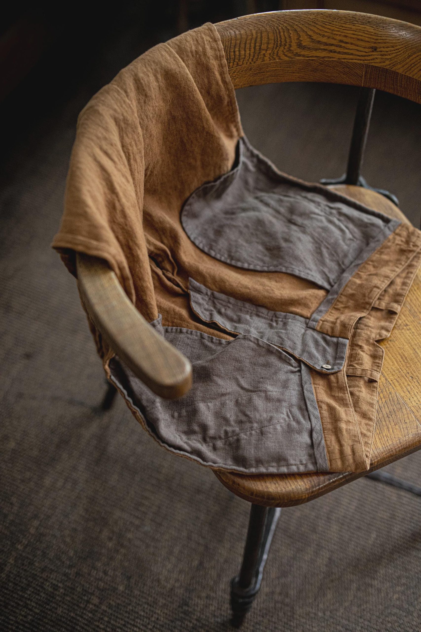 Brown linen trousers laid open on a curved wooden chair, angled close-up of the waist; belt loops, gray lining pieces with stitched borders, overlapping front seam, small white button on a gray flap, creases across fabric, and chair armrest visible.