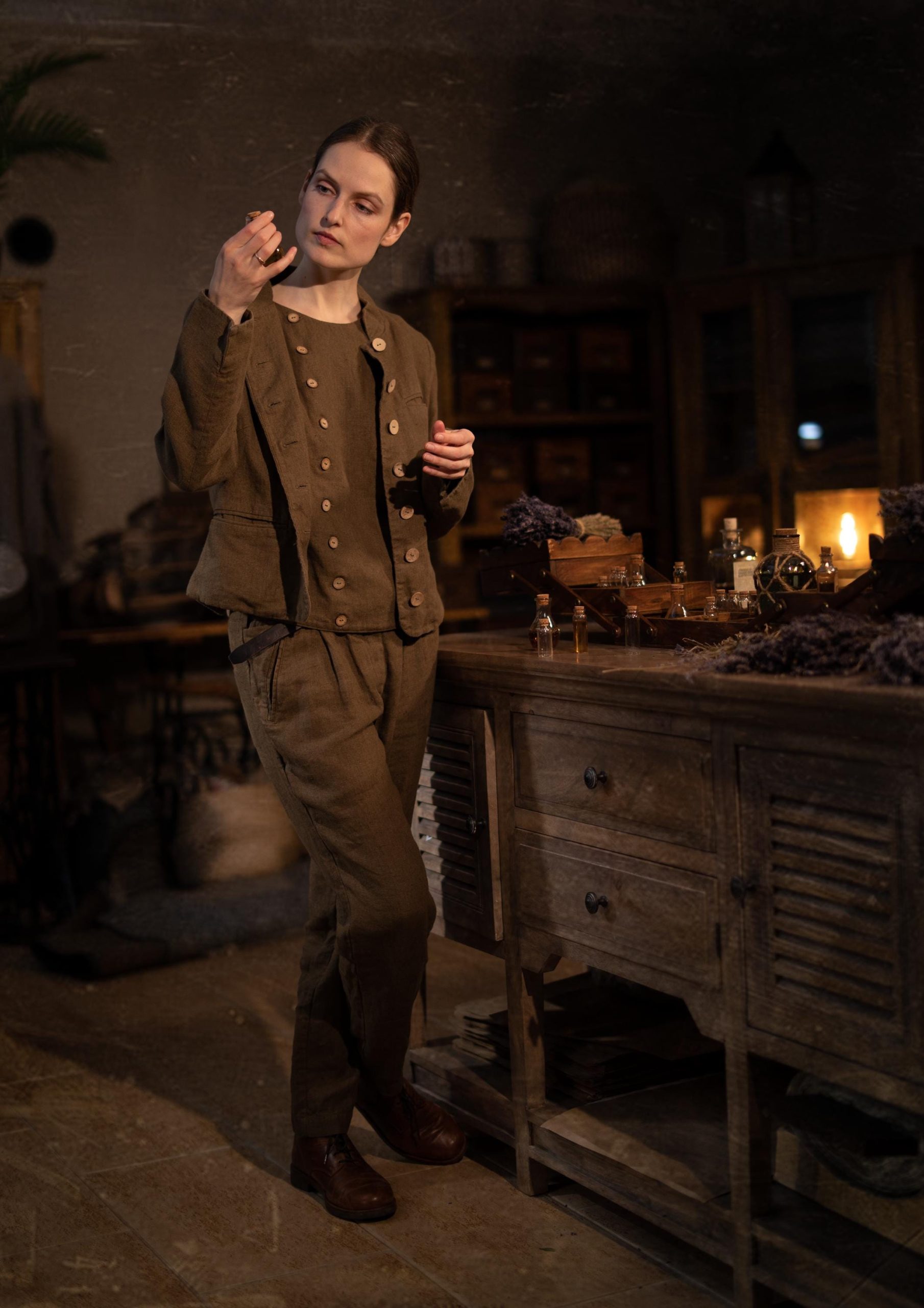 Brown linen trousers on a model, full-length front/side view; pleat folds at waist, angled pocket opening, side seam line, wrinkles at knee, narrow ankles above brown shoes; drawers and bottles on a sideboard.