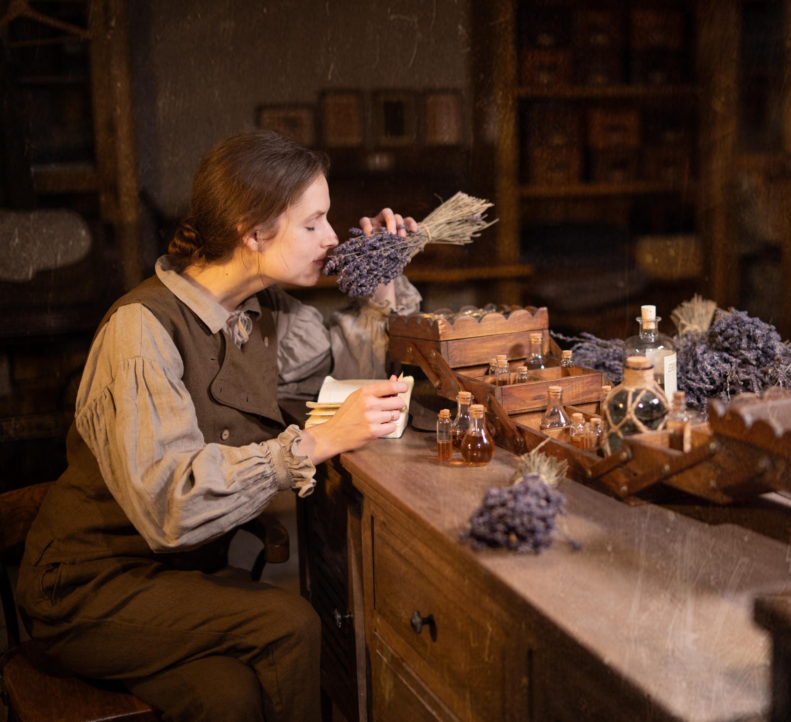 Brown linen trousers on a model seated at a wooden desk, side crop waist-to-knee; waistband hidden, angled hip pocket, folds at the seat, creased thigh, bent knee, and side seam line; lavender and small glass bottles on the desk.