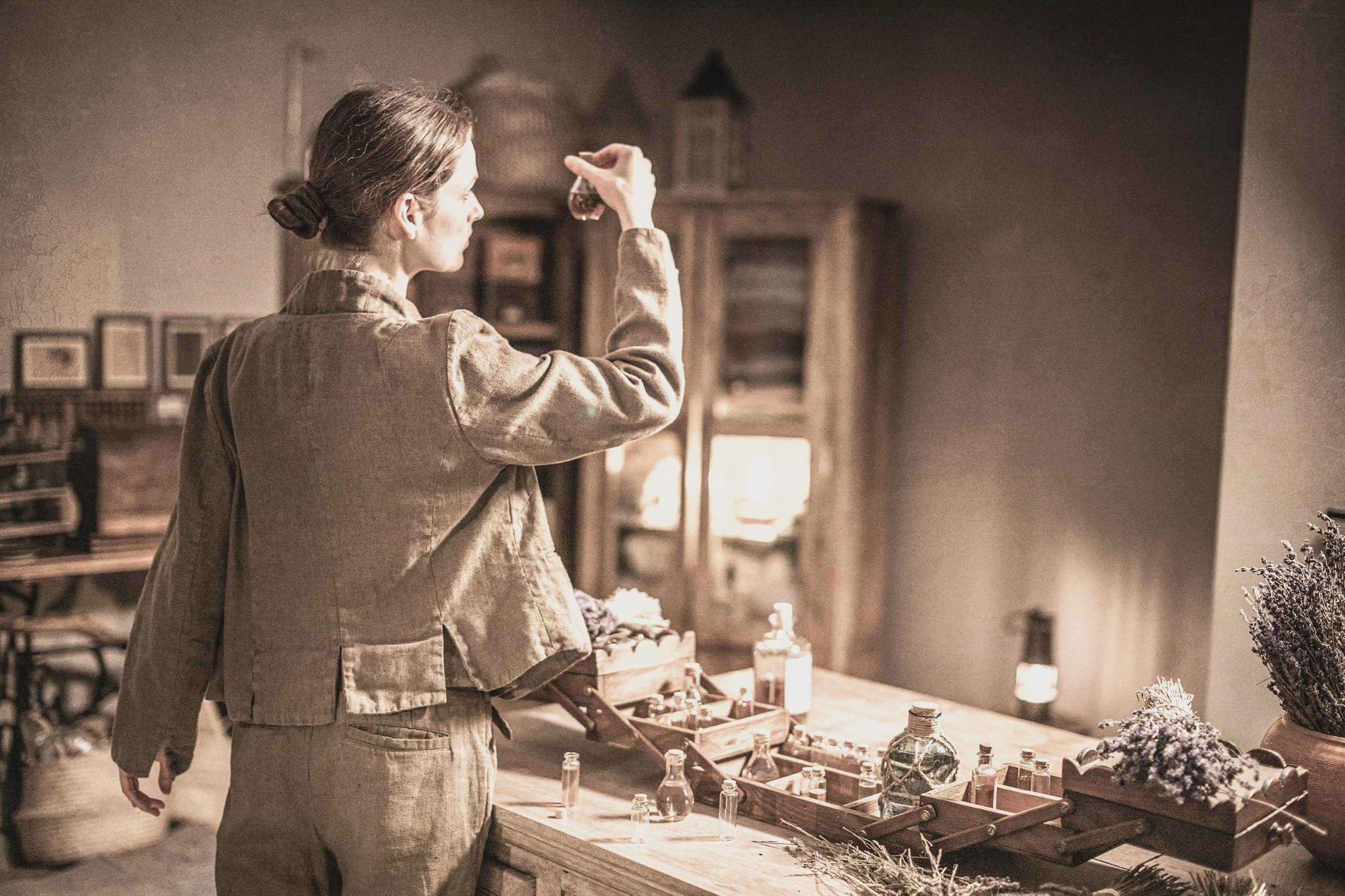Linen pants worn by a model, full-length back view; waistband covered by a short jacket, one back pocket, center back seam, straight legs, and wrinkled linen texture; she holds up a small glass bottle over a table of jars.