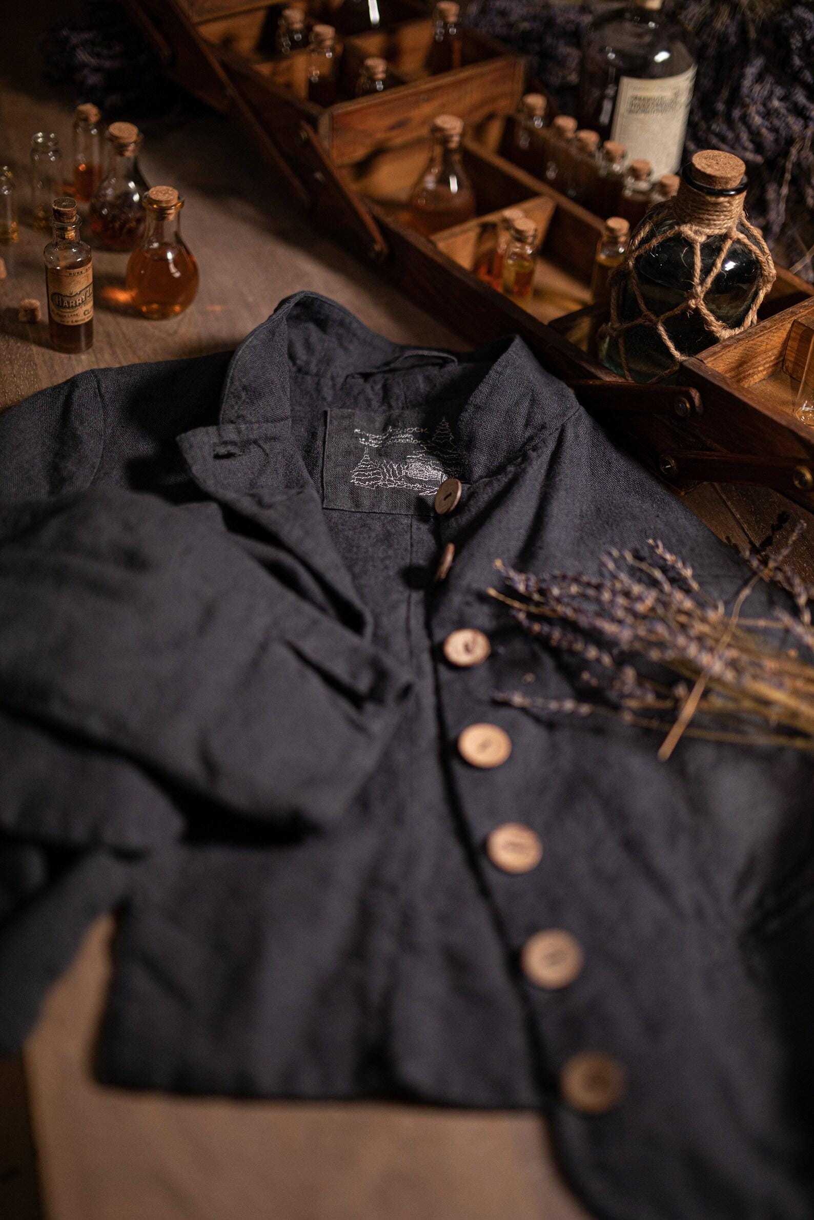Charcoal linen jacket laid on a table, top-down crop; stand collar, button front with light round buttons, long sleeve folded across, visible buttonholes, small stitched emblem inside neck area, and wrinkled texture; lavender stems and glass bottles nearby.