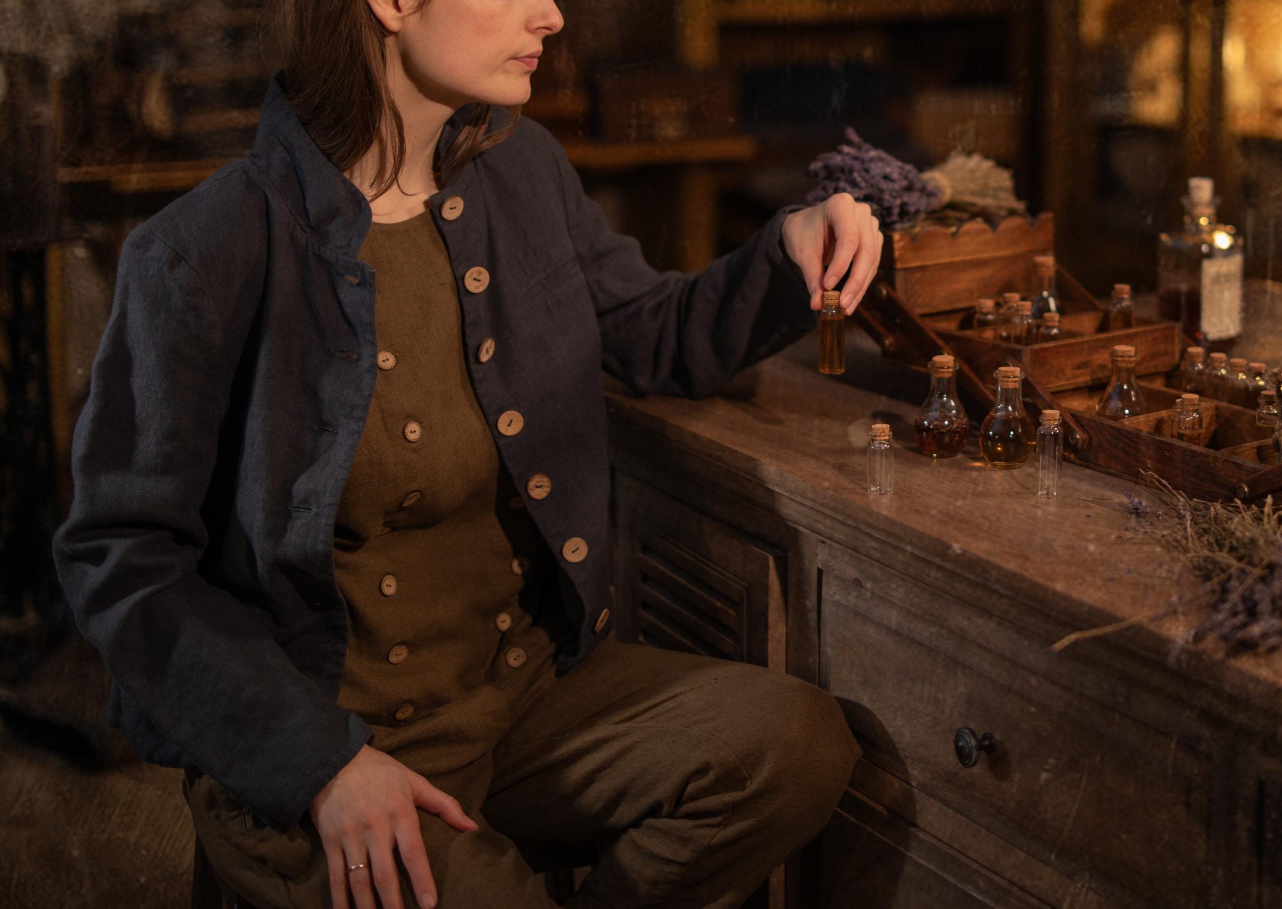 Charcoal linen jacket worn on a model, cropped chest-to-hip view; short stand collar, row of light buttons down front, long sleeves, open front showing brown buttoned top, and wrinkled texture; hand holds a small bottle above a tray of tiny jars and lavender.