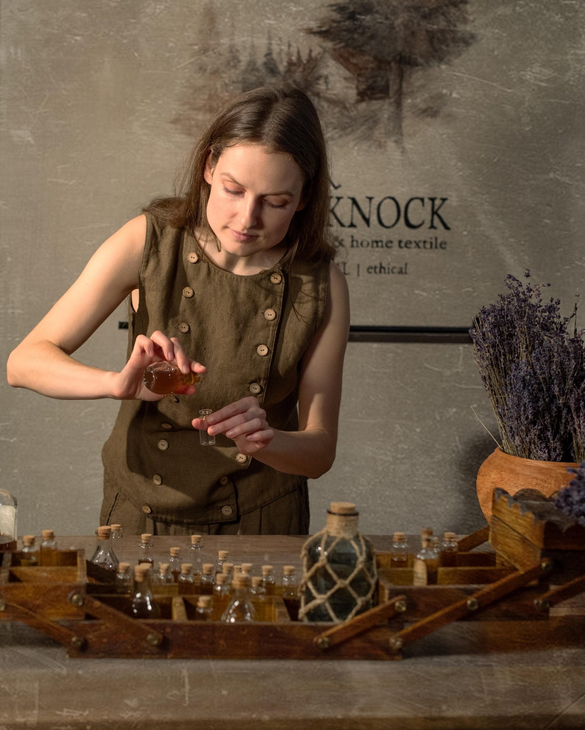 Brown linen waistcoat on model, waist-up; sleeveless with round neck, light buttons along both front edges, layered front panels; she fills a small vial at a table of glass bottles, including a rope-net bottle and scattered purple lavender.