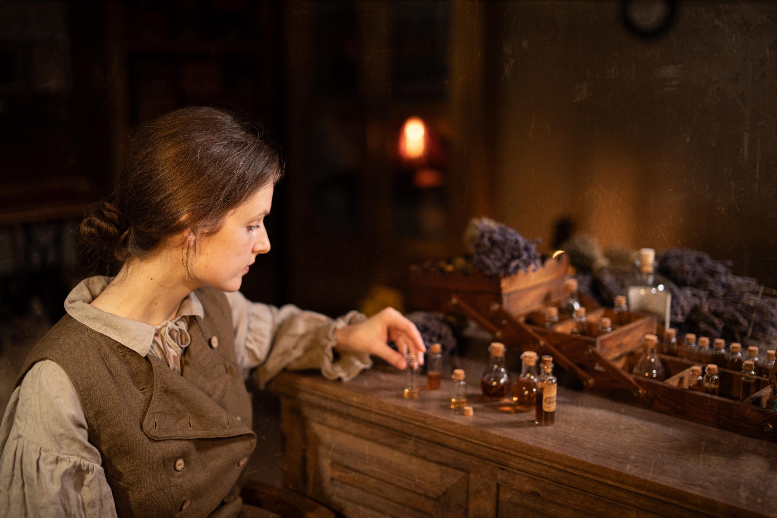 Brown linen vest on seated model, side-profile waist-up; worn over light gray blouse with ruffled collar and tie; front has wide flap with buttonholes and small buttons; her hand reaches toward corked glass vials on a wooden cabinet.
