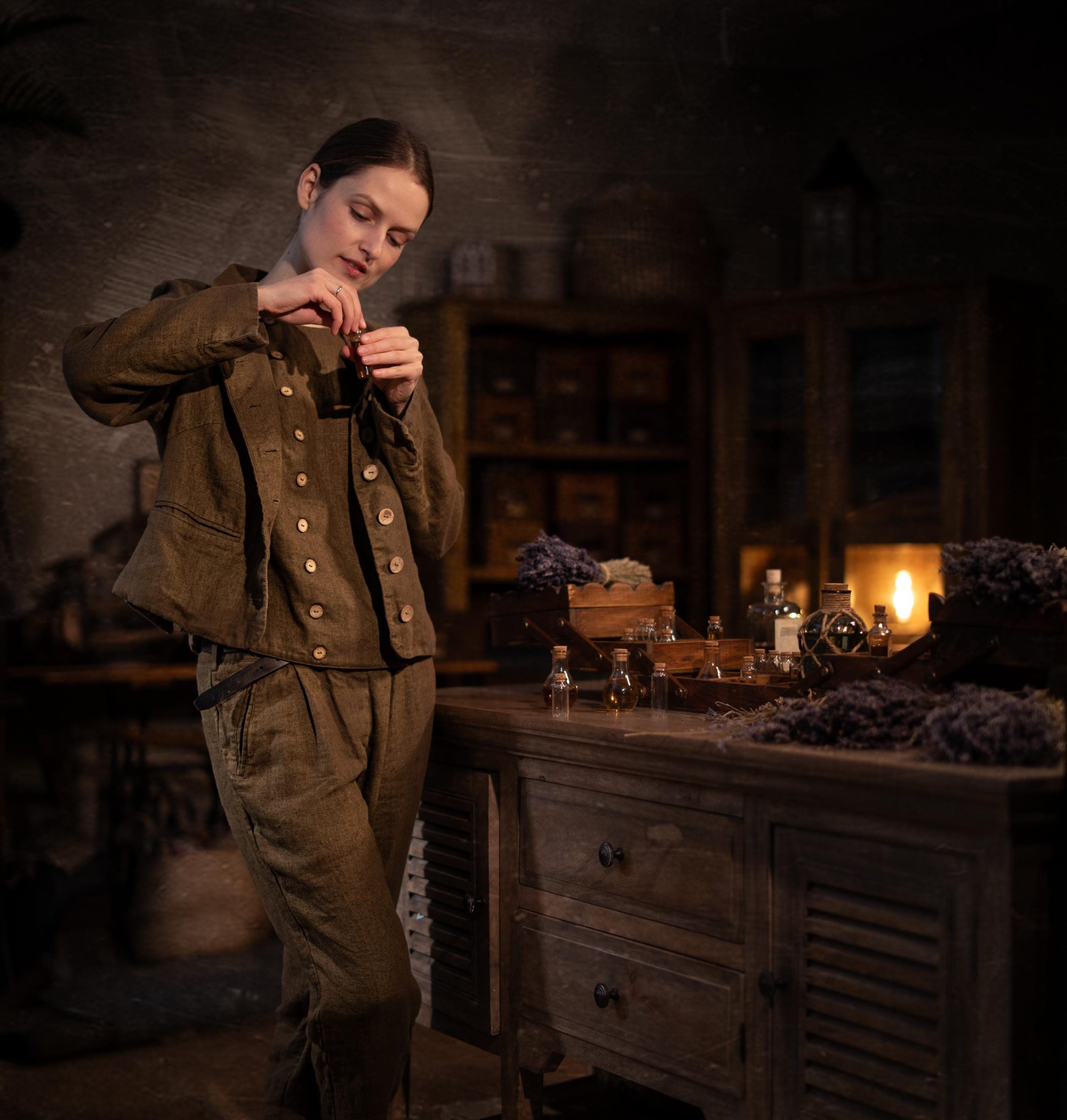 Brown linen jacket worn by a model, three-quarter front view; round neck, button front with light buttons, long sleeves, visible front seam lines, pocket opening at hip, hem at hip, wrinkled linen texture; wooden drawers, bottles, candle behind.