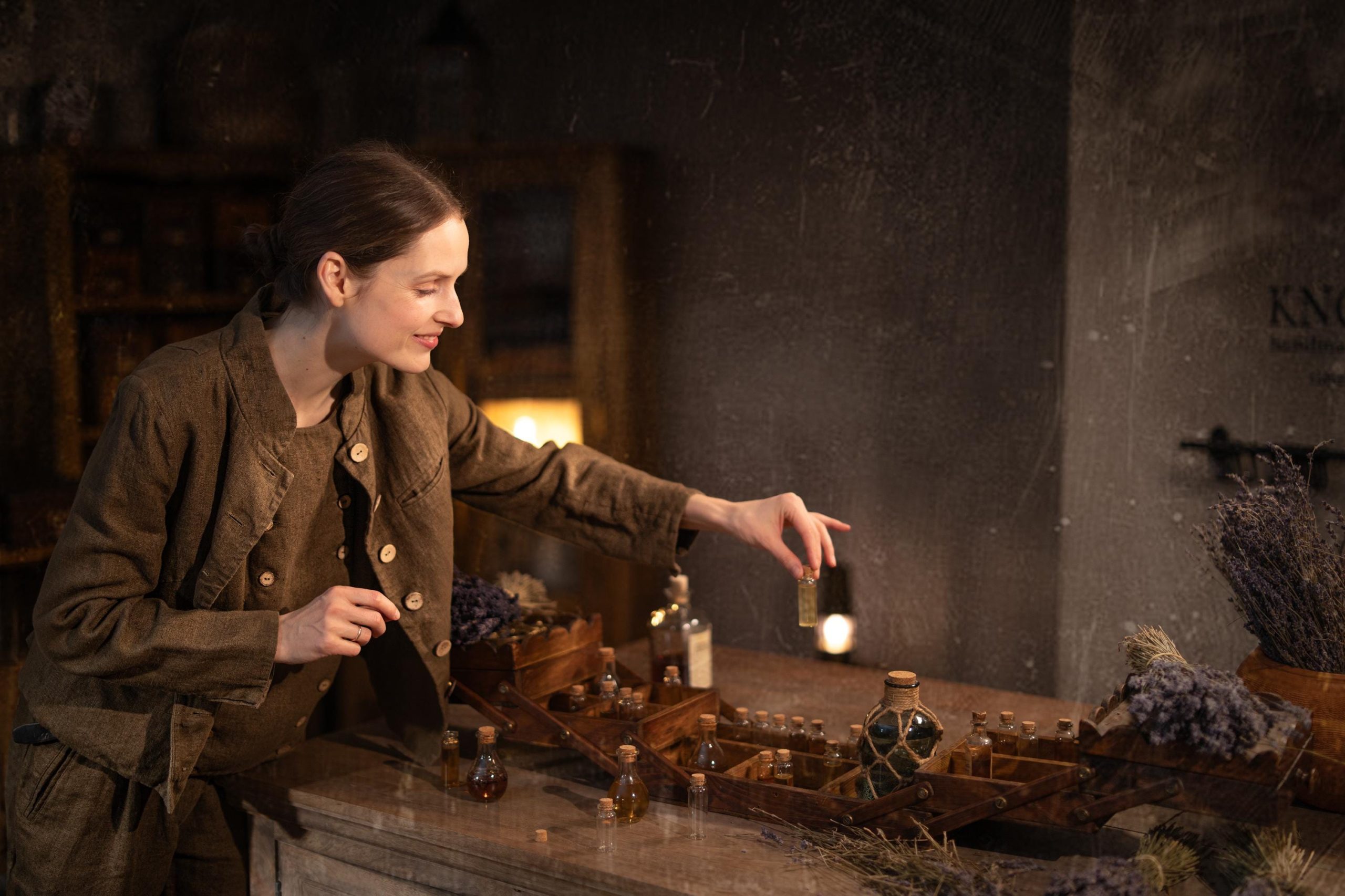 Brown linen jacket worn by a model, waist-up side/three-quarter view; collarless neckline, light buttons down the front, long sleeves, straight cropped hem, and visible stitching at the front; wooden tray of small bottles and dried flowers on table.