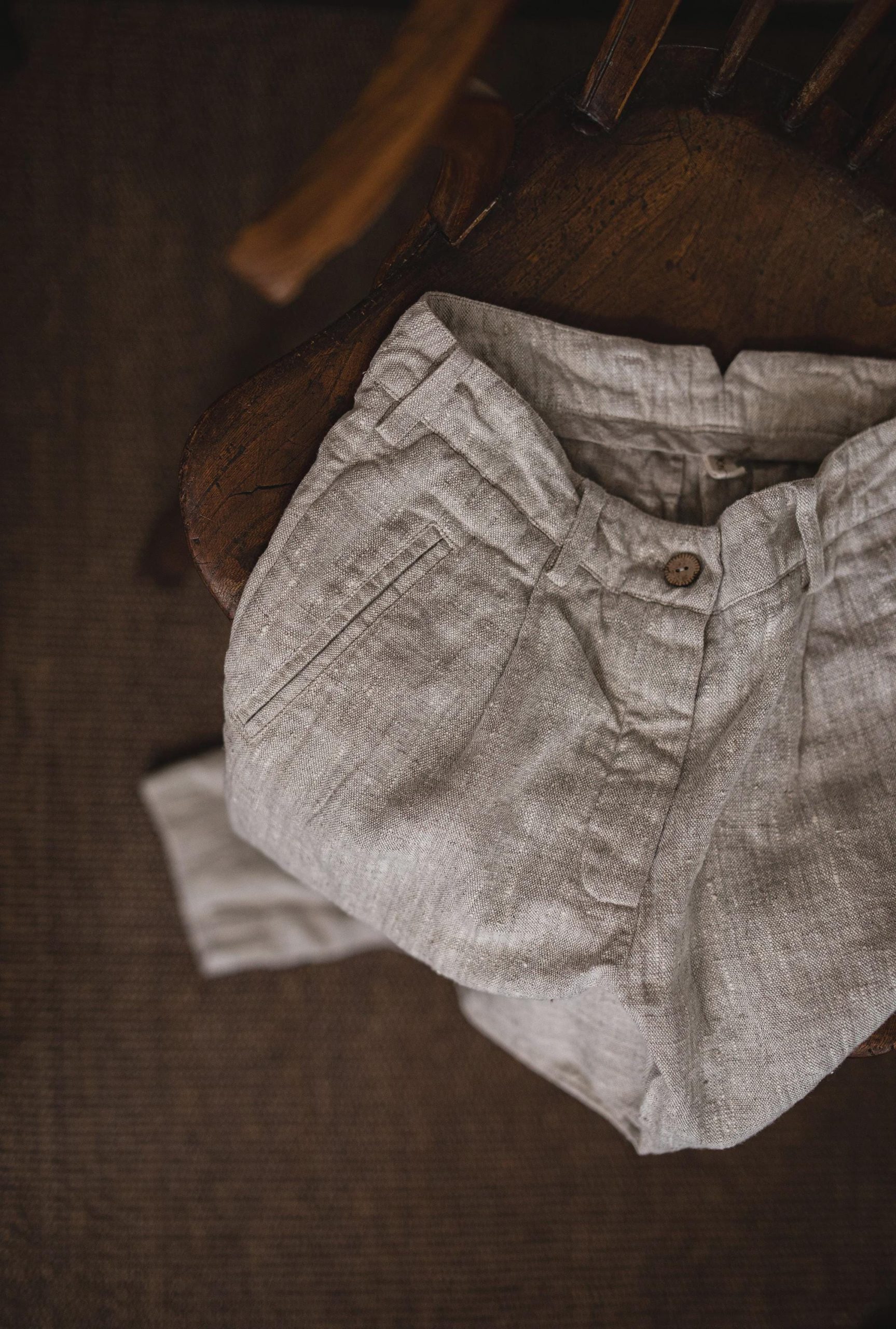 Natural flax linen pants folded on a wooden chair, top-down waist detail; waistband with belt loops, single brown button, overlapping front placket, stitched pleat folds, narrow pocket opening with topstitching, and wrinkled linen texture.