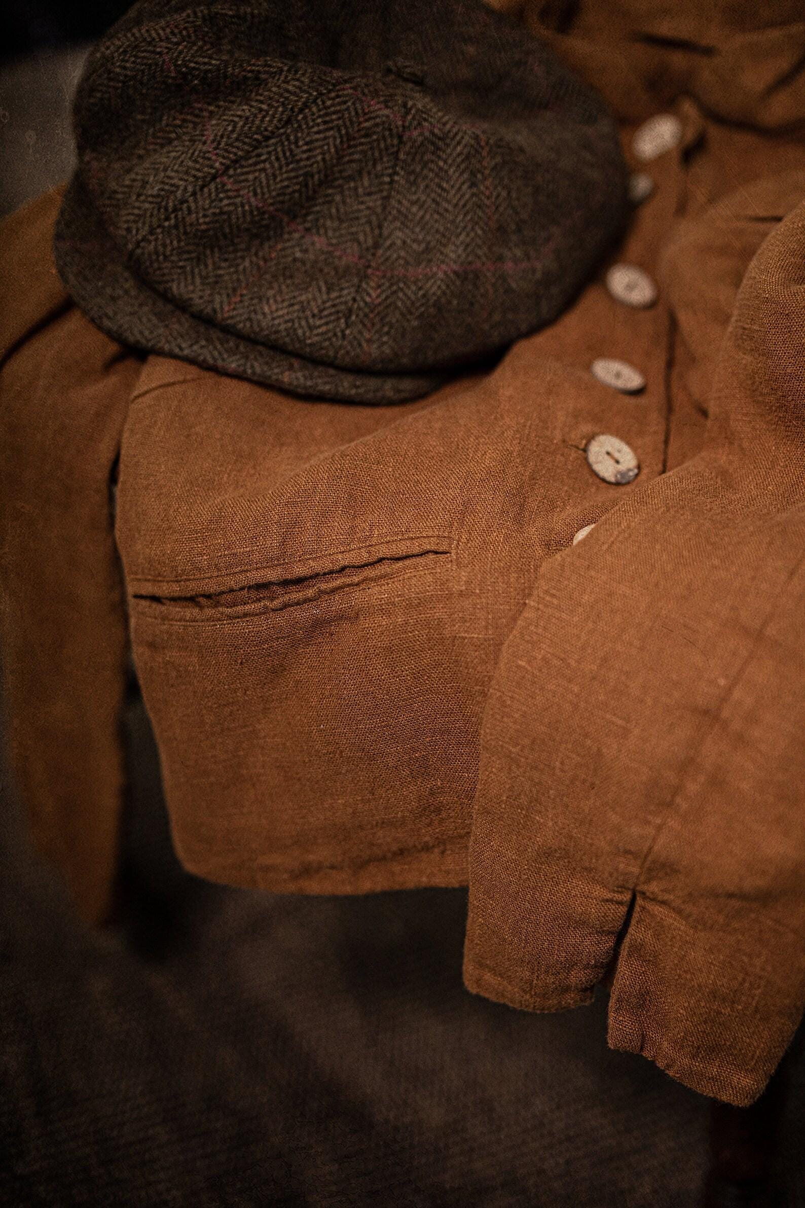 Linen jacket in a flat lay close-up; light round buttons on the front, welt pocket opening, sleeve folded at the cuff, visible seam stitching and creased texture, with a dark gray herringbone cap resting on top.