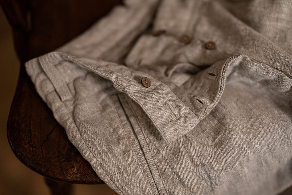 Folded melange linen trousers draped over a dark wooden chair, showing the waistband, buttons, and front seams.